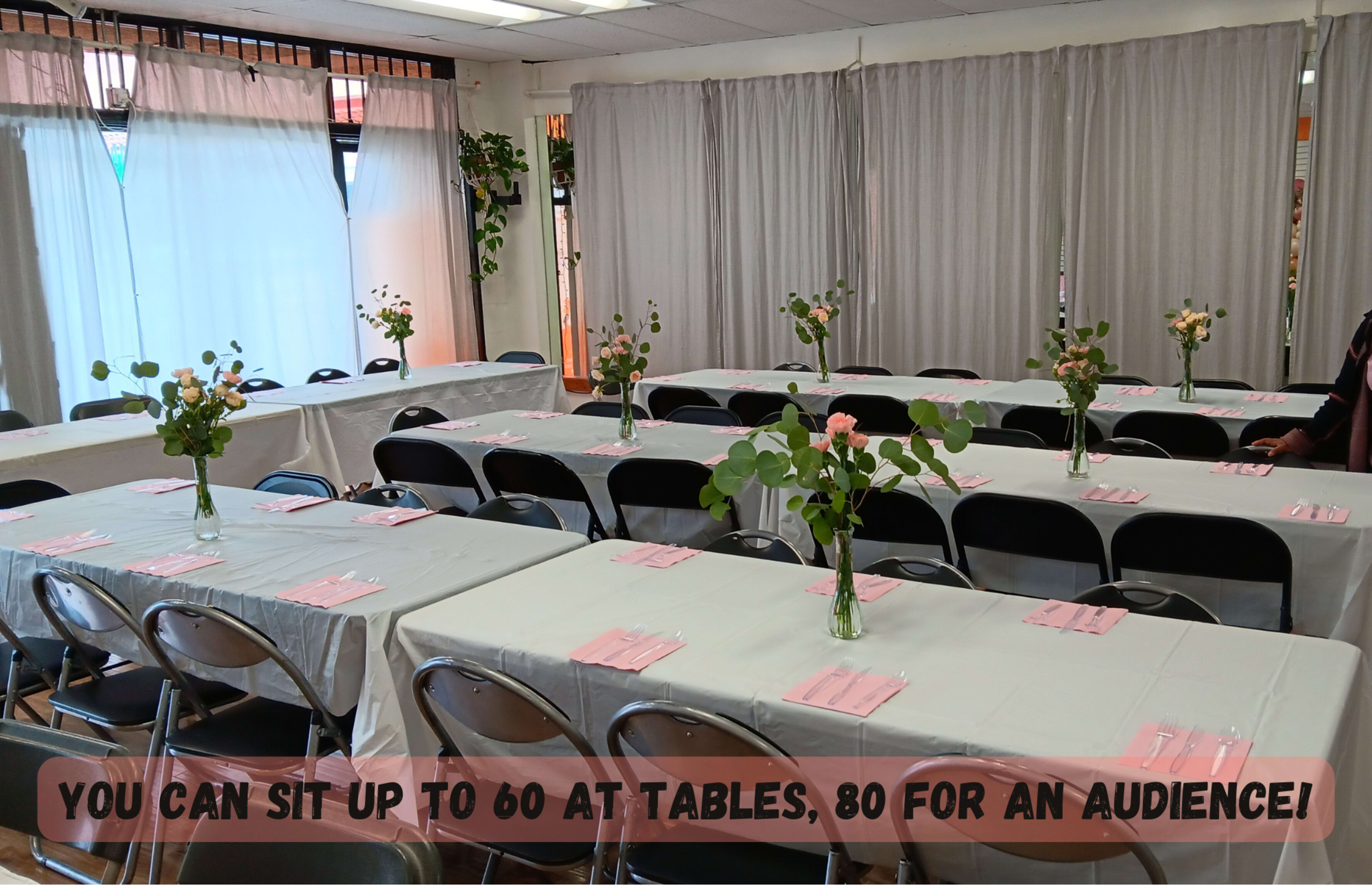 A spacious room is set up with multiple tables covered in white linens and decorated with flower arrangements, ready to accommodate guests for an event.