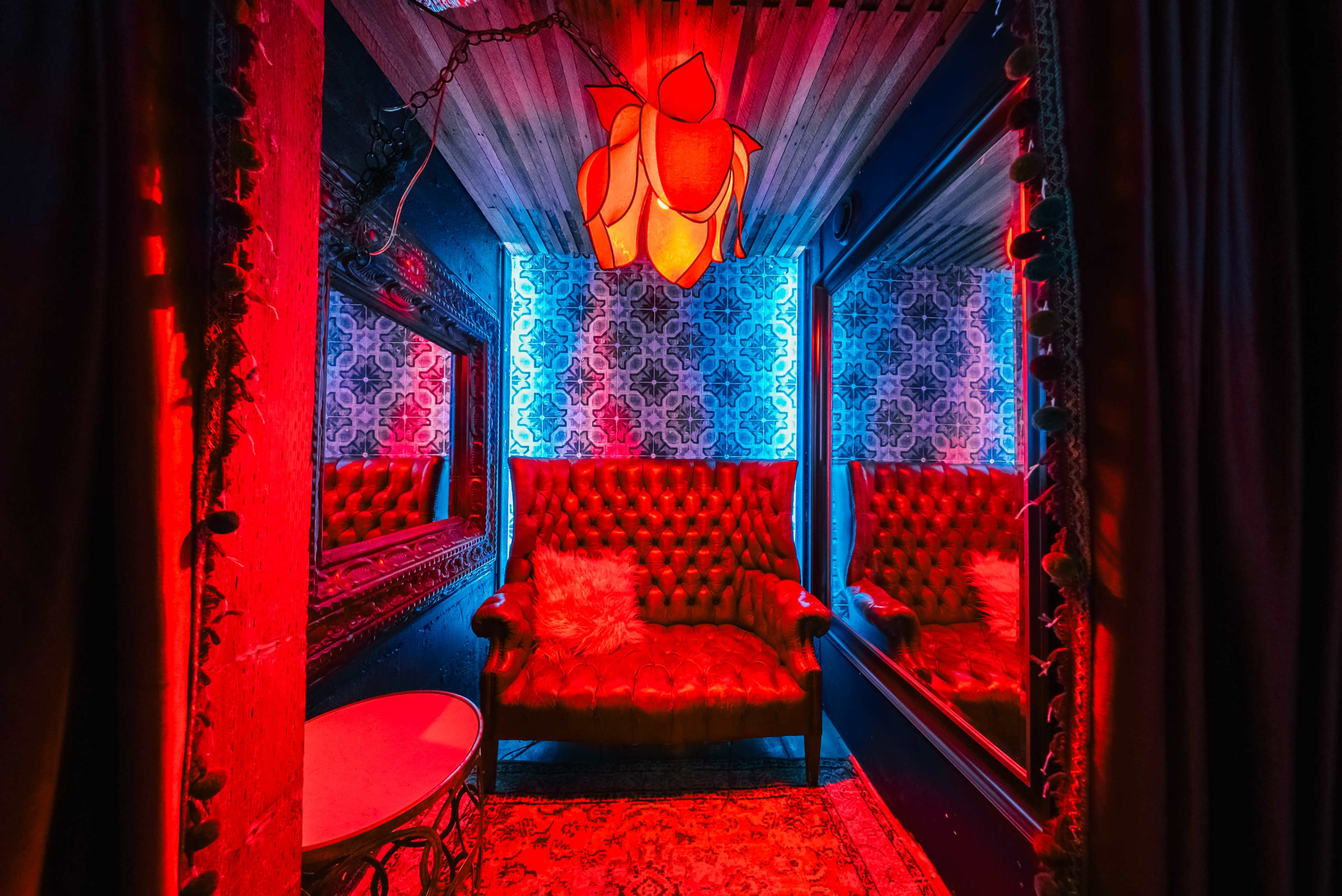 A dimly lit lounge features a red and blue color scheme with a tufted red sofa, a round table, and decorative mirrors.