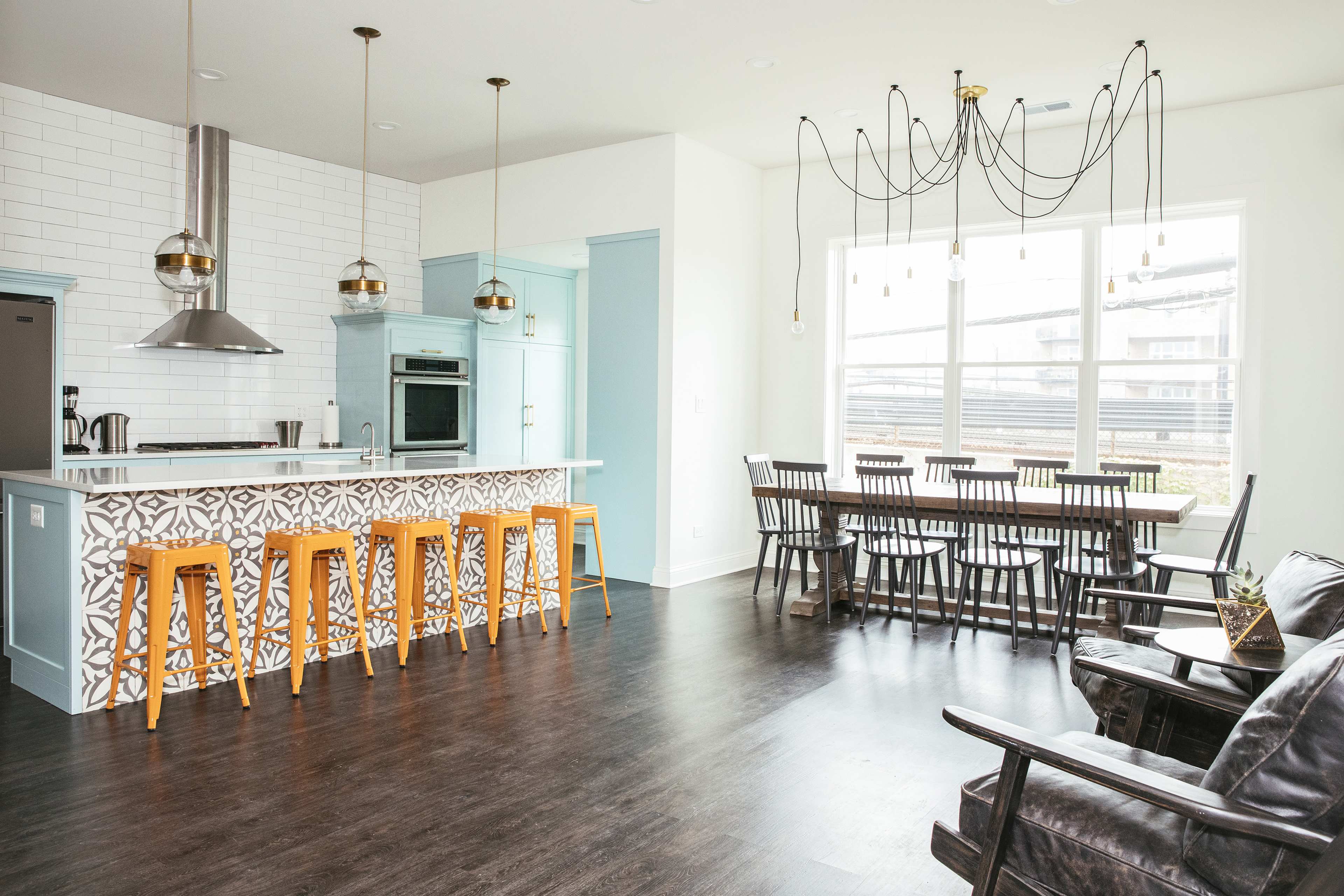A modern kitchen and dining area with a bar featuring wooden stools, a large dining table, and a stylish light fixture above.
