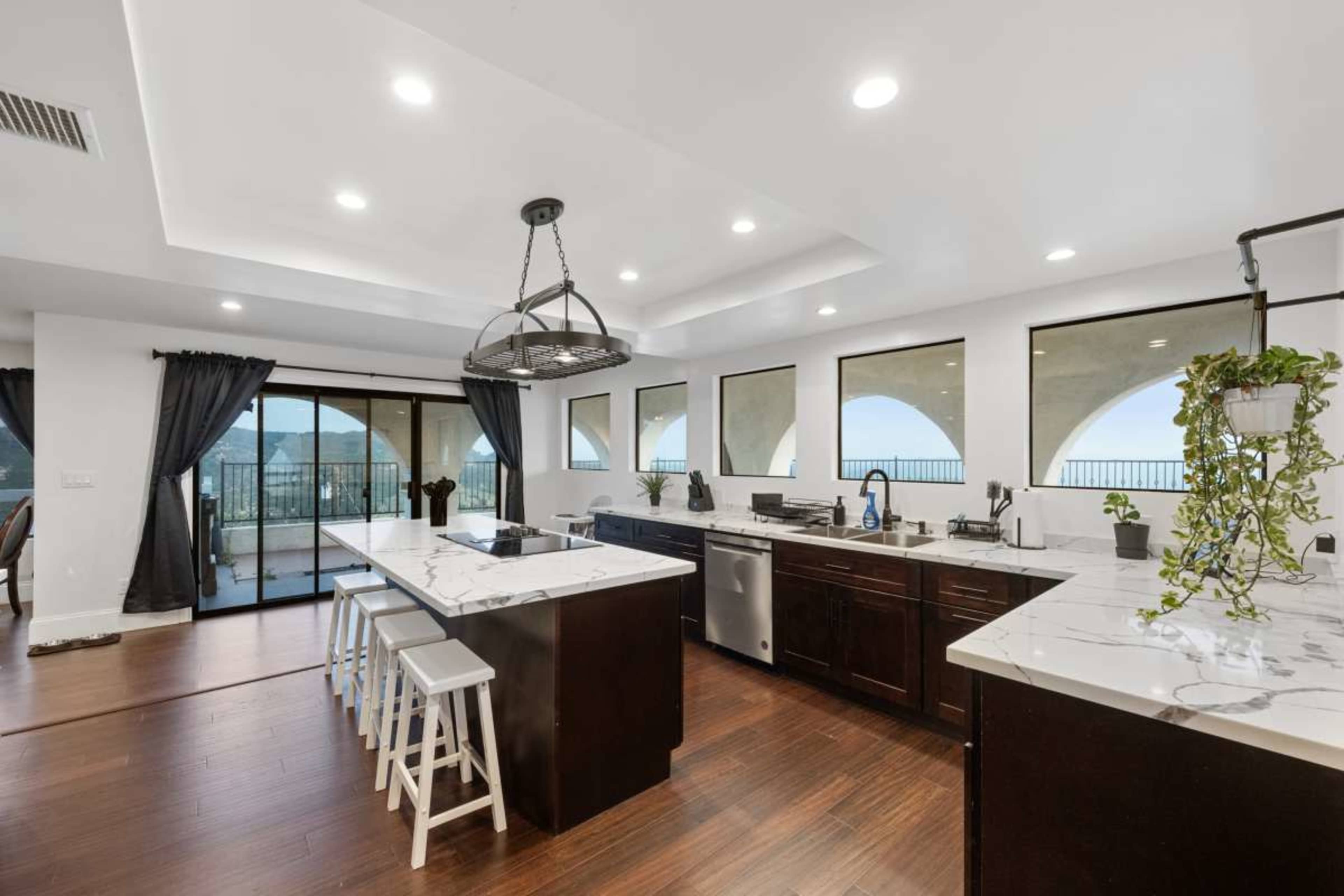 The image shows a modern kitchen with a large central island, dark cabinetry, and multiple windows providing a view of the outdoors.