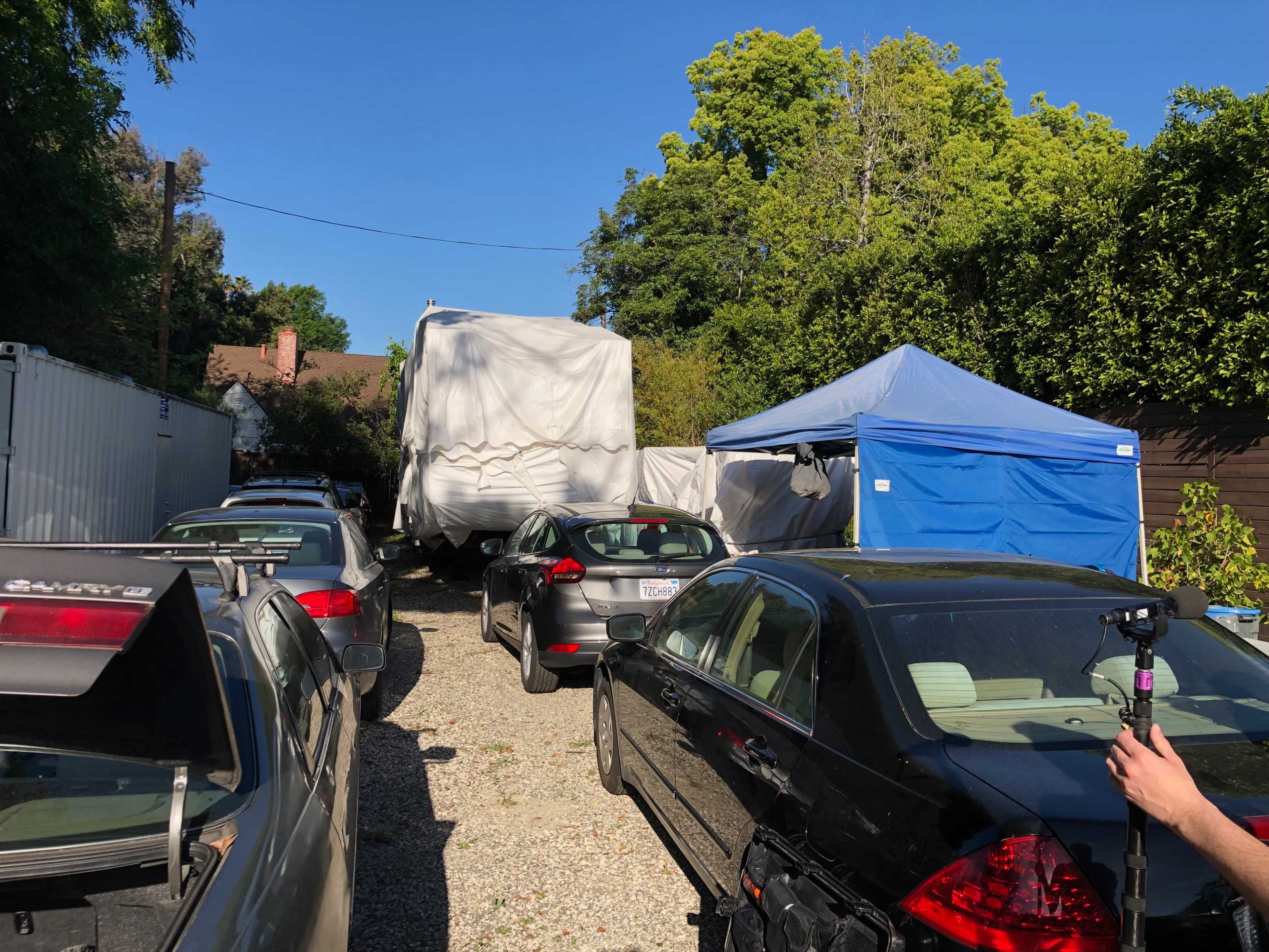 A narrow driveway is lined with parked cars, leading to a large covered trailer and two tents set up in a backyard area.