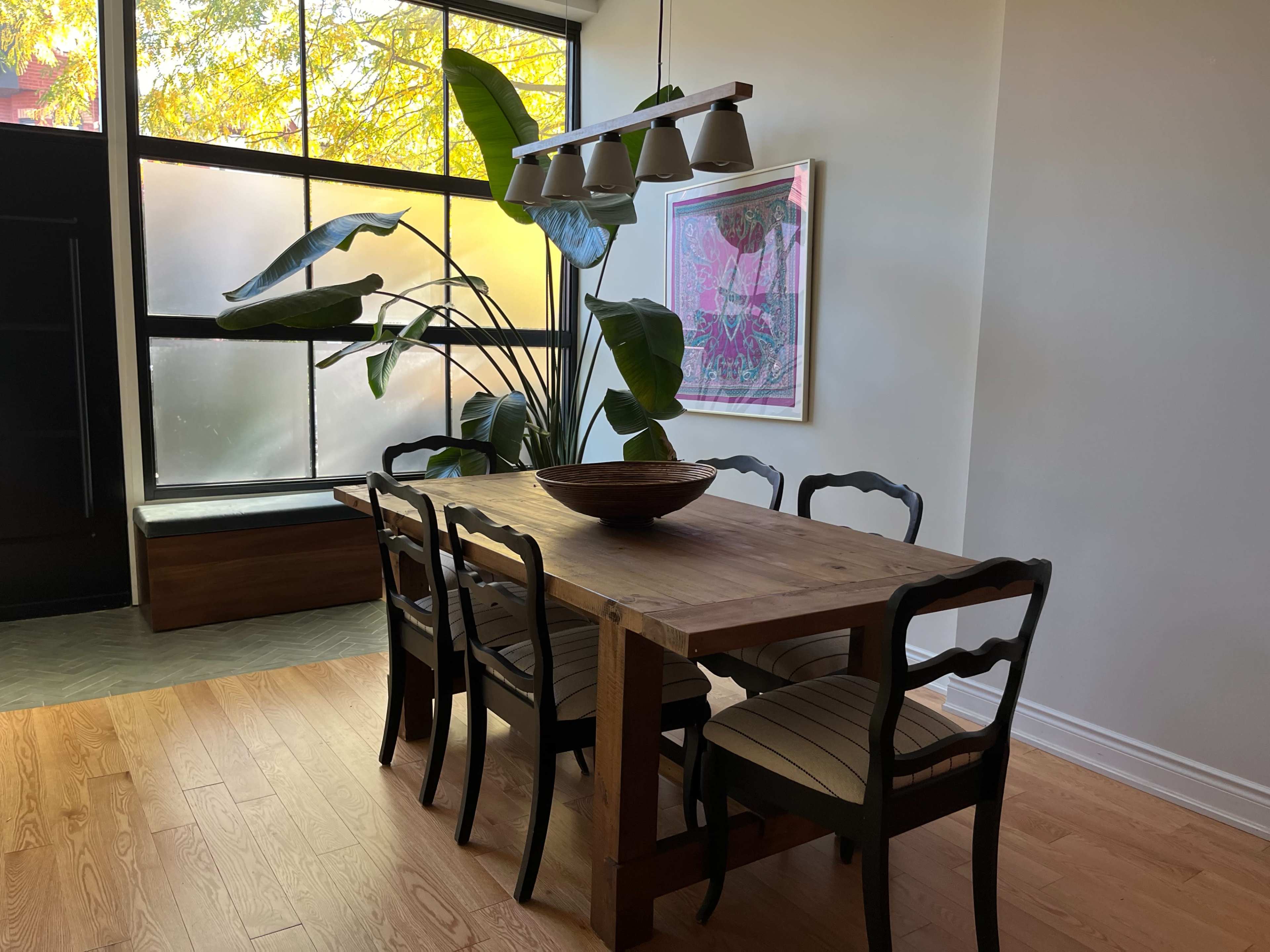 A dining table with six chairs is positioned in a well-lit room featuring large windows and a potted plant nearby.