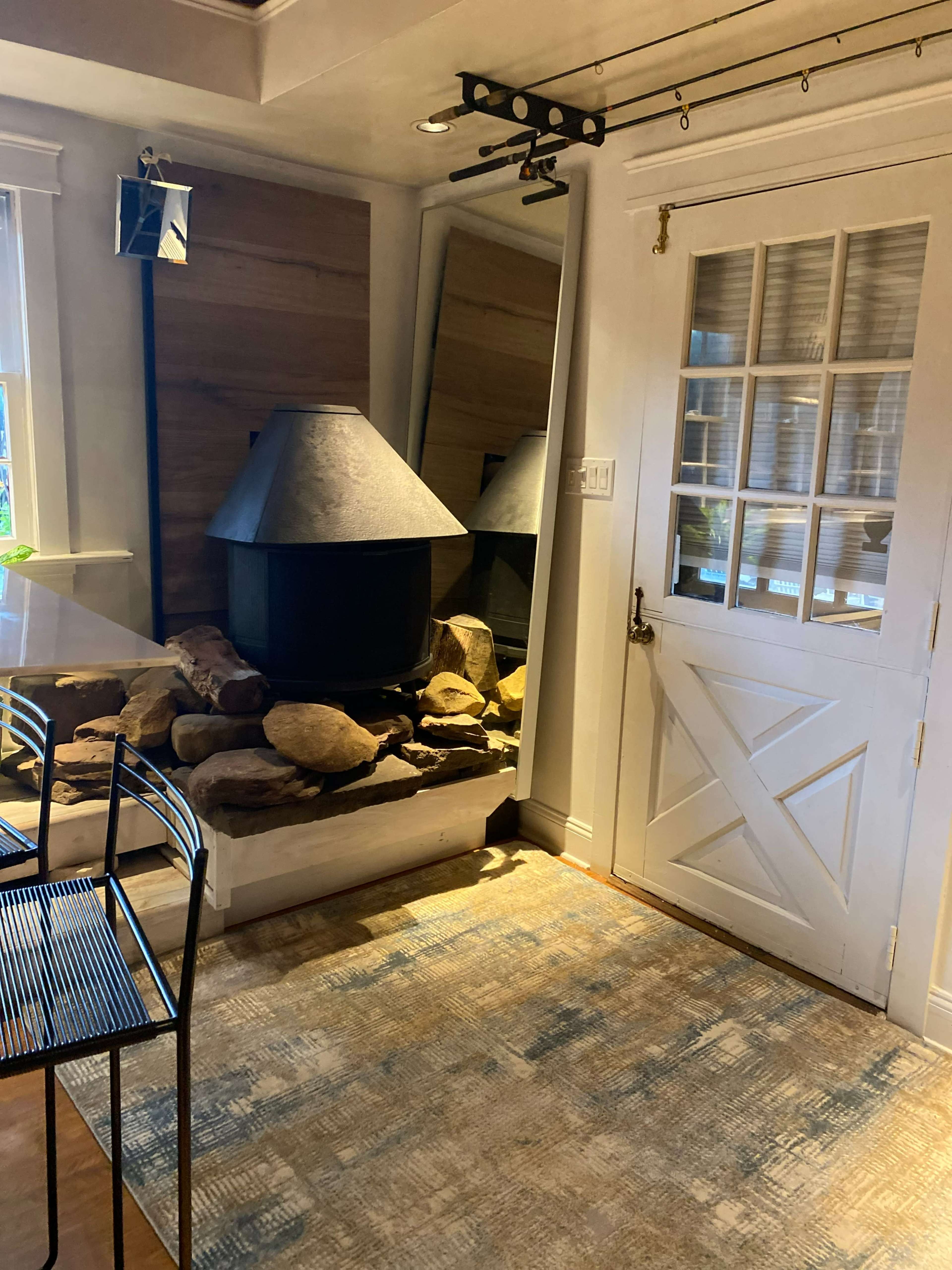 A cozy interior space featuring a large, black fireplace surrounded by stones, a mirrored wall, a door with window panes, and a patterned rug on the floor.