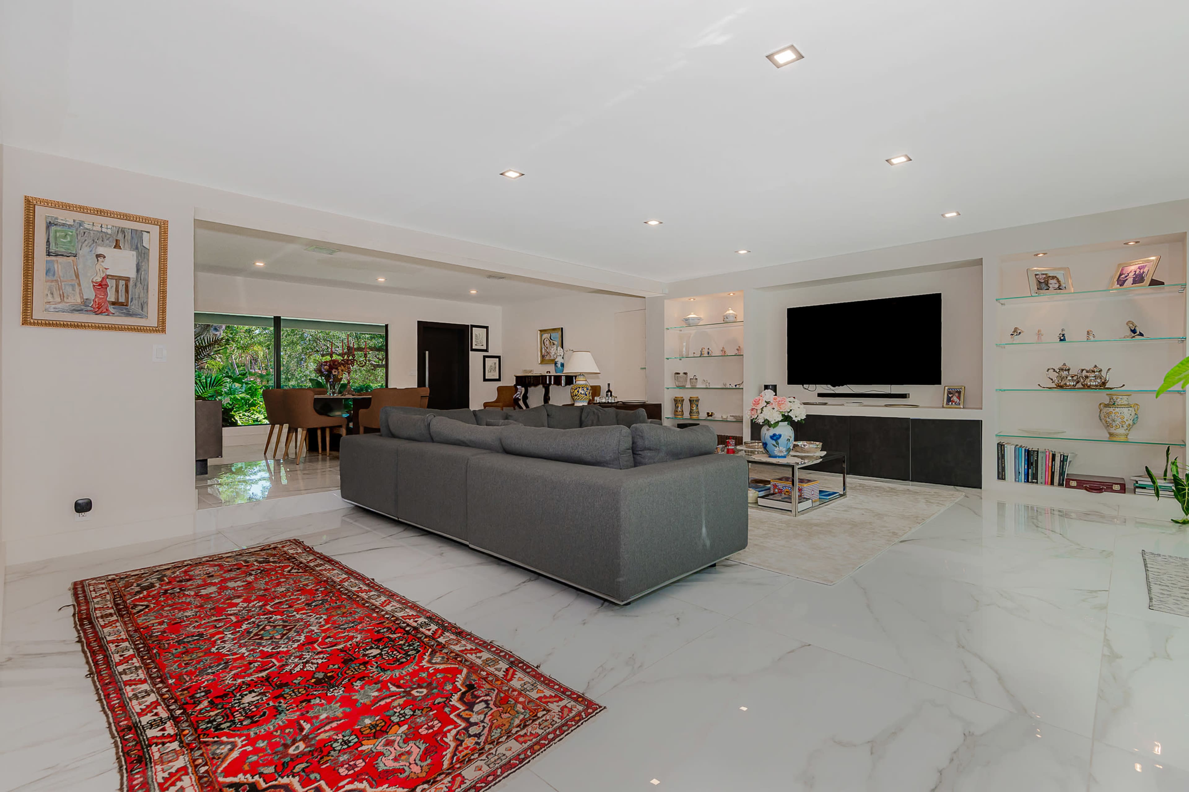 A modern living room features a grey sectional sofa, a large television mounted on a wall unit, and a plush area rug against glossy marble flooring.