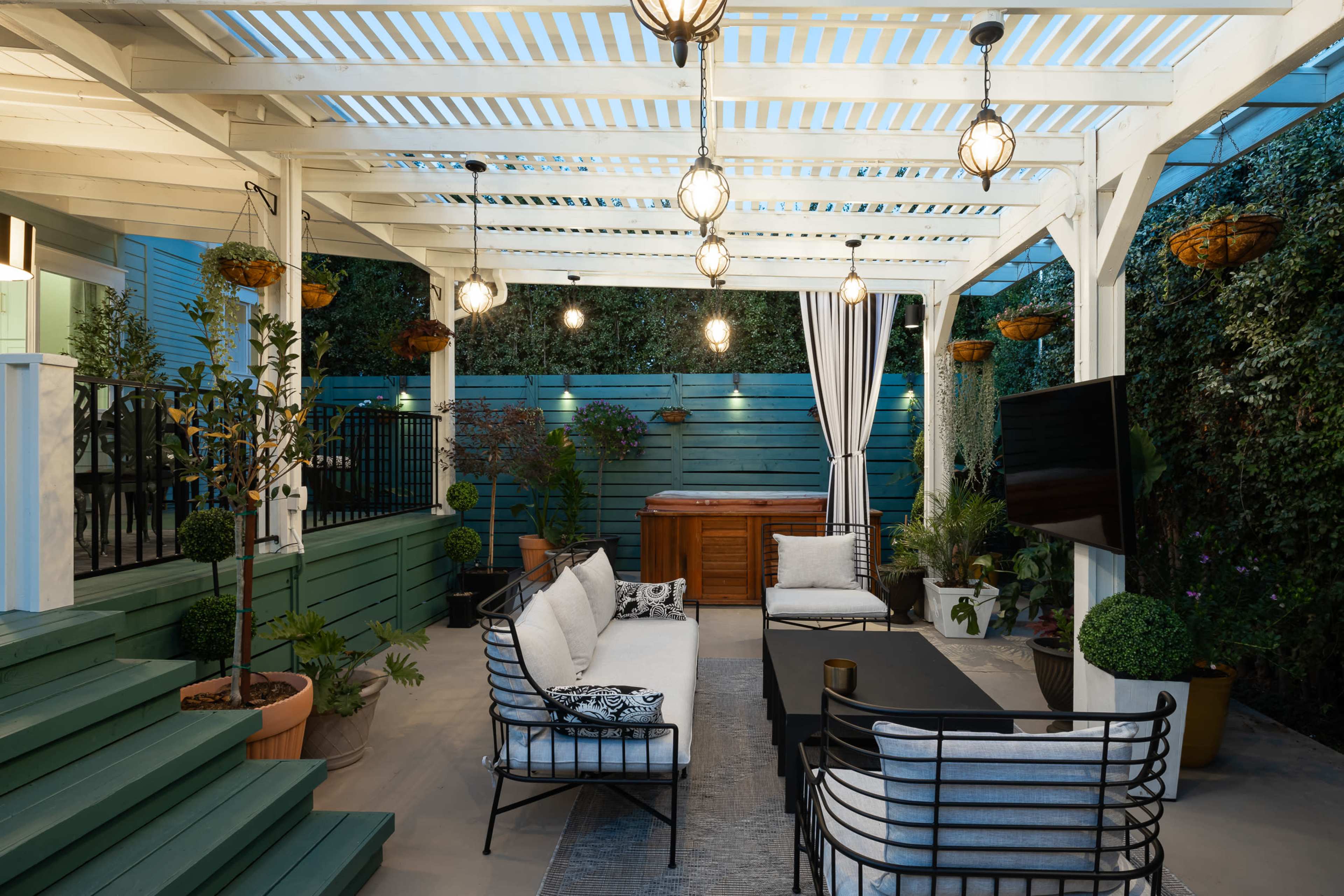 A covered patio features a seating area with a sectional sofa, a coffee table, and a television, surrounded by decorative plants and lighting.