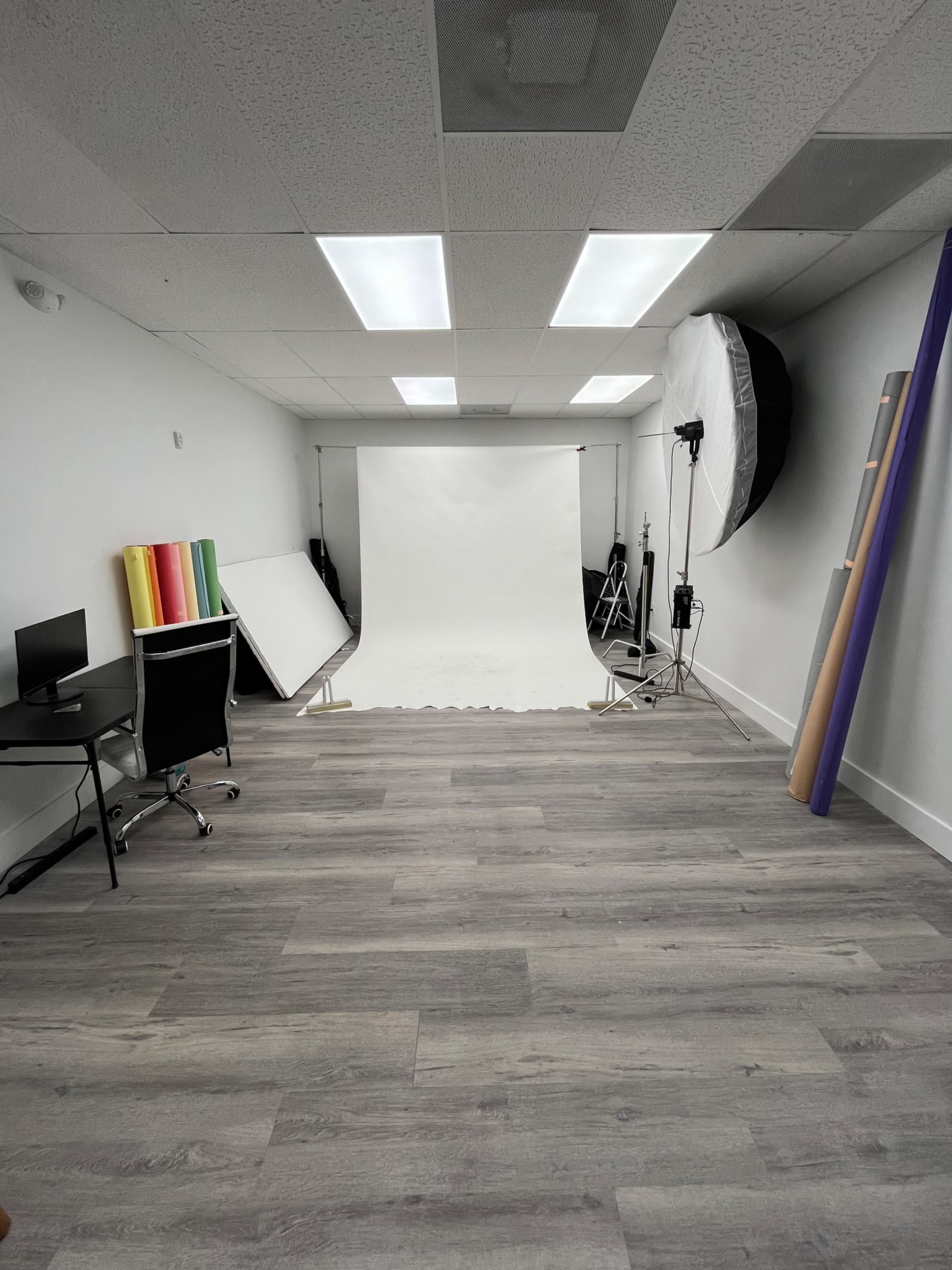 The image shows a photography studio with a white backdrop, various colored rolls of paper, a light setup, and a desk with a chair.