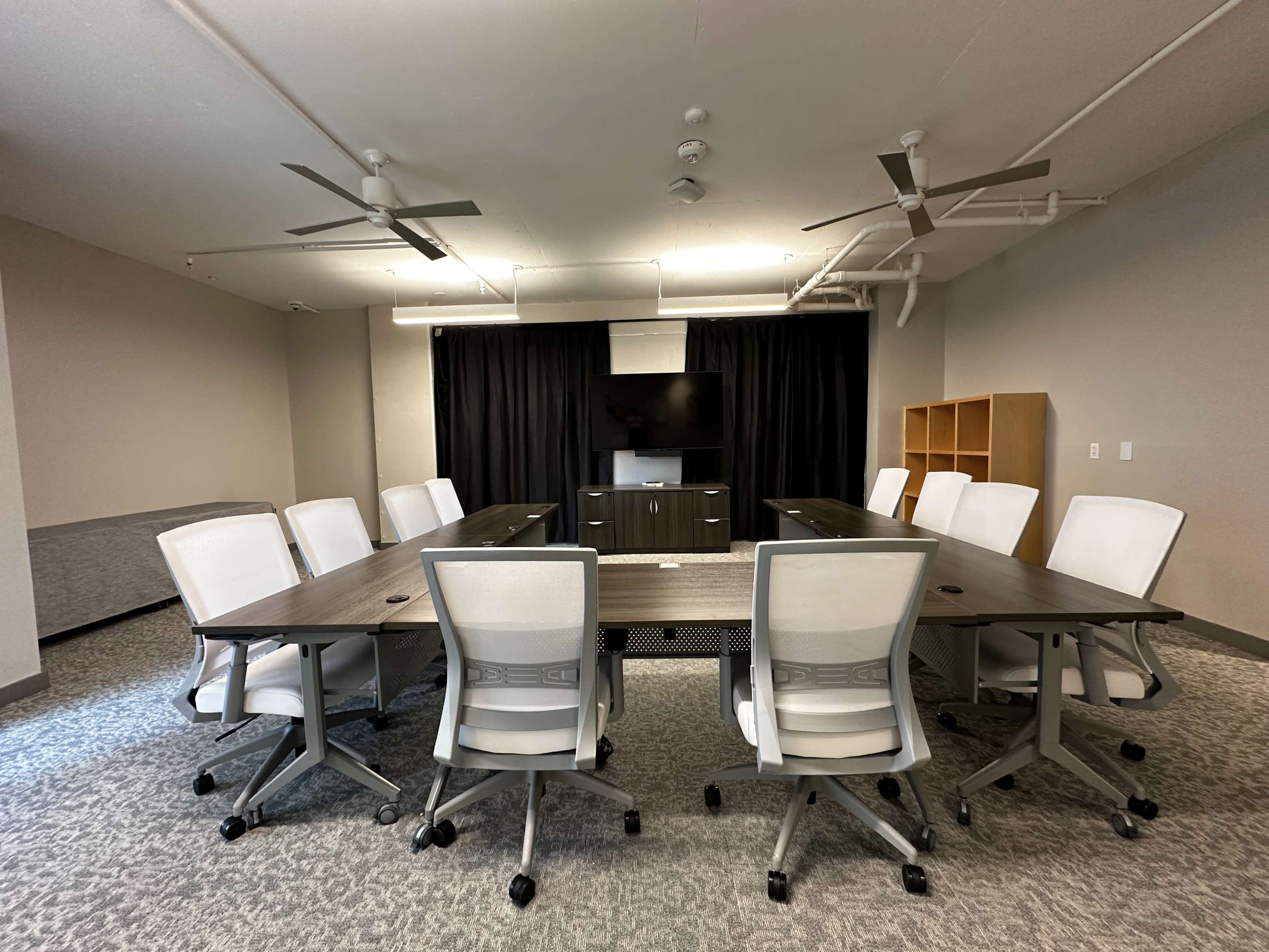 The image shows a modern conference room with a large table surrounded by white rolling chairs, a television on a stand at one end, and black curtains at the front.