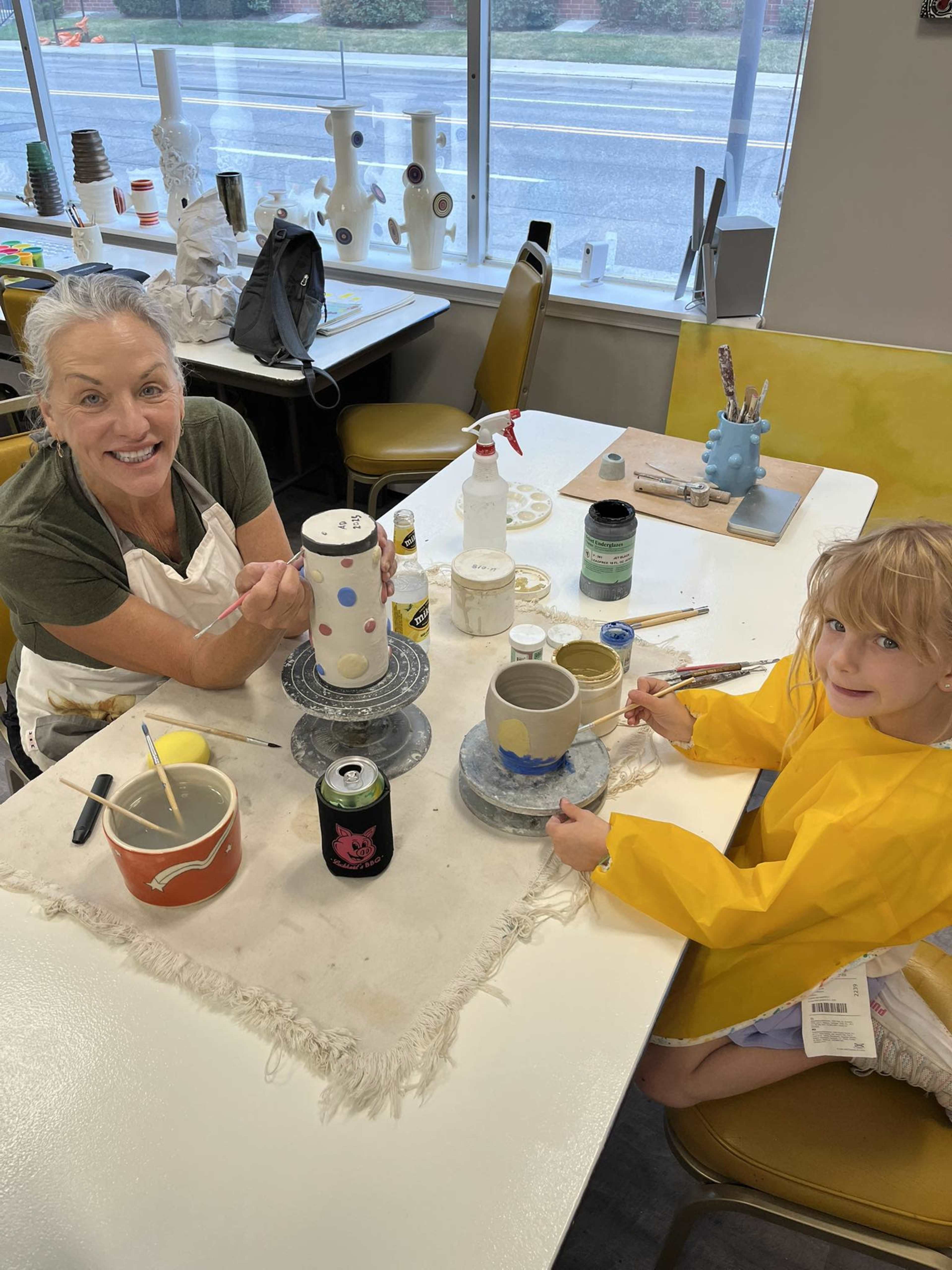 A woman and a young girl are crafting at a table, painting pottery pieces in a brightly lit studio.