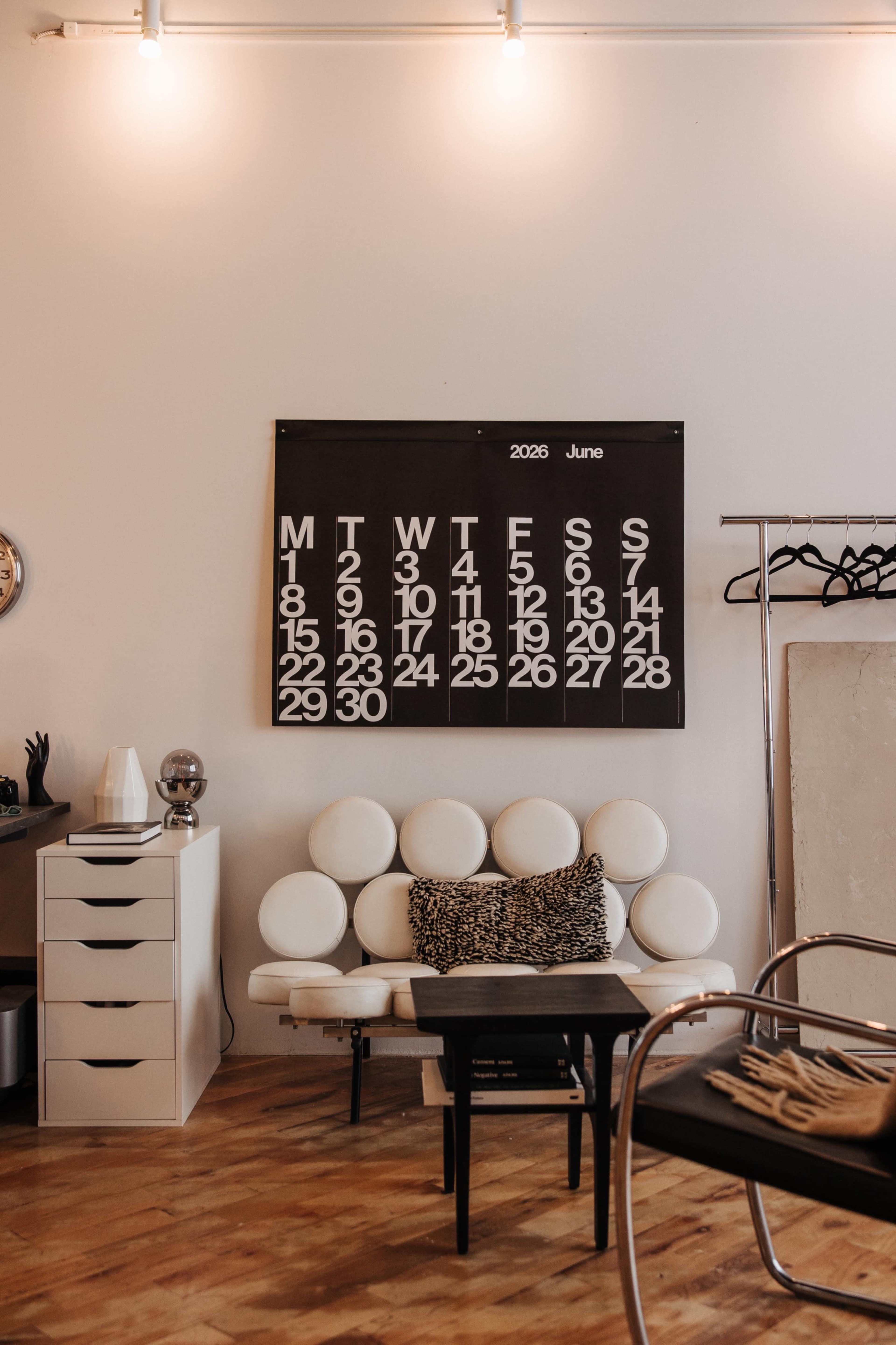 The image shows a minimalist room featuring a white sofa with round cushions, a calendar for June 2028 on the wall, and a small black coffee table.