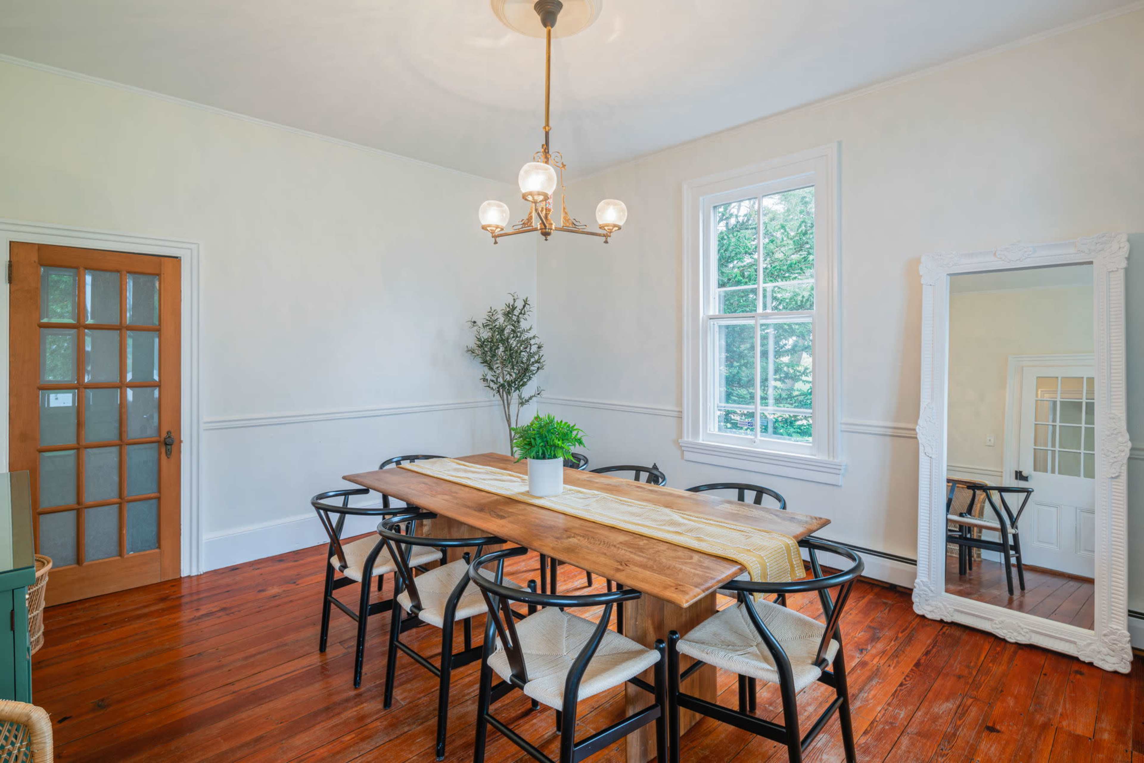 A dining room features a long wooden table surrounded by black chairs, with a chandelier overhead and large windows allowing natural light.