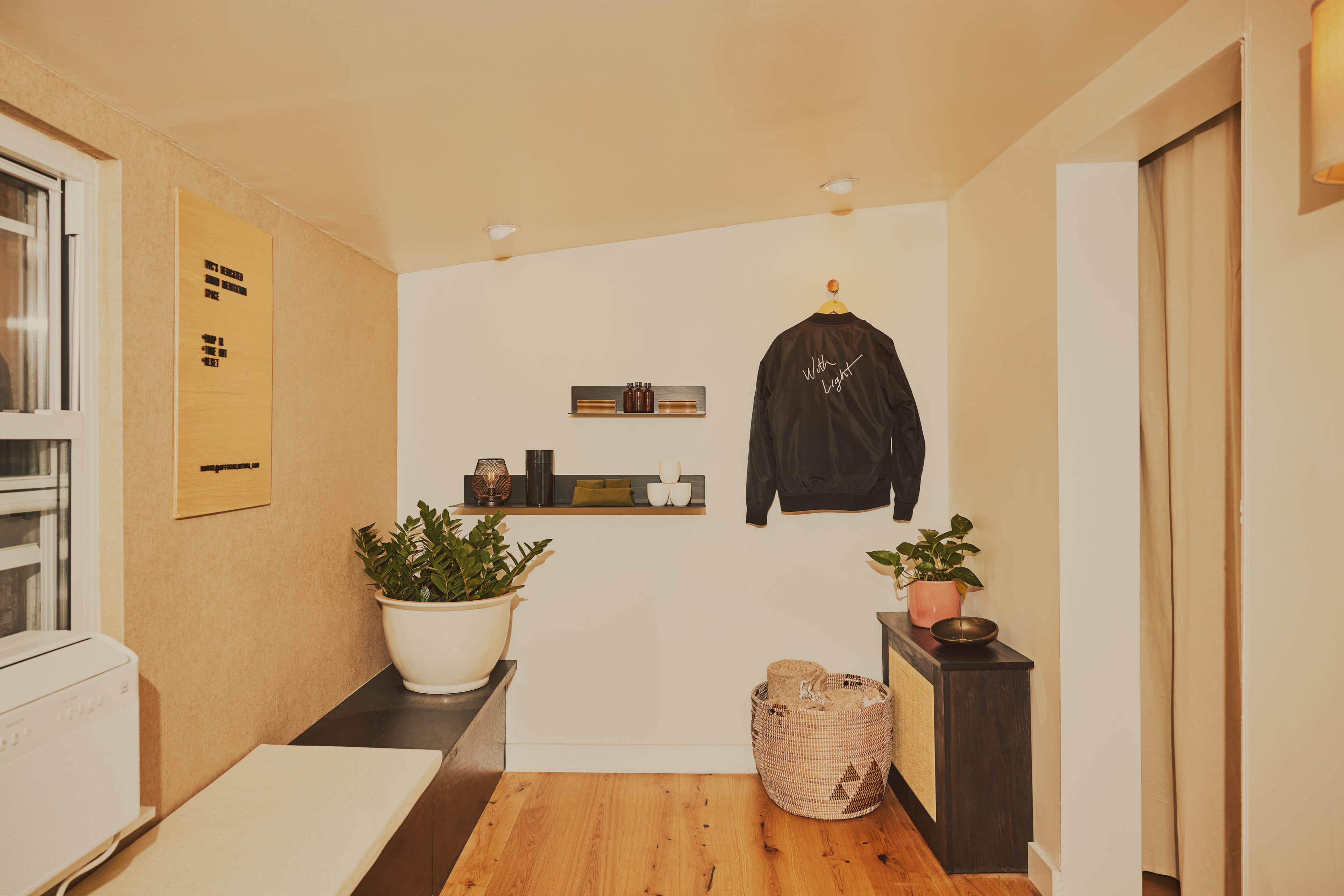 The image shows a cozy corner of a room with light-colored walls, a small shelf displaying decor, a plant, a black jacket hanging, and a seating area with a bench and a woven basket.