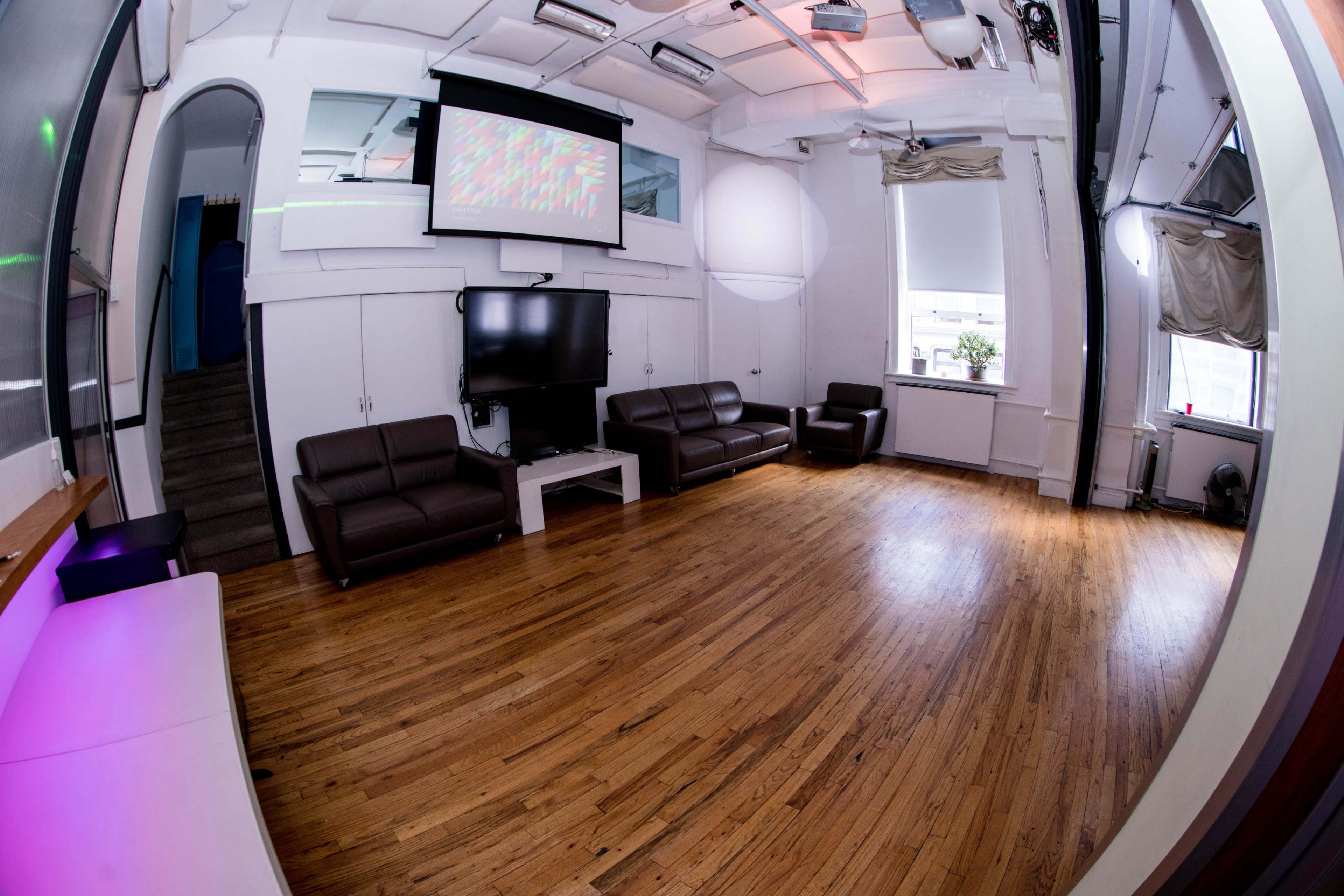 The image shows a spacious room with wooden flooring, two brown leather sofas, a mounted television, and a projection screen on the wall.