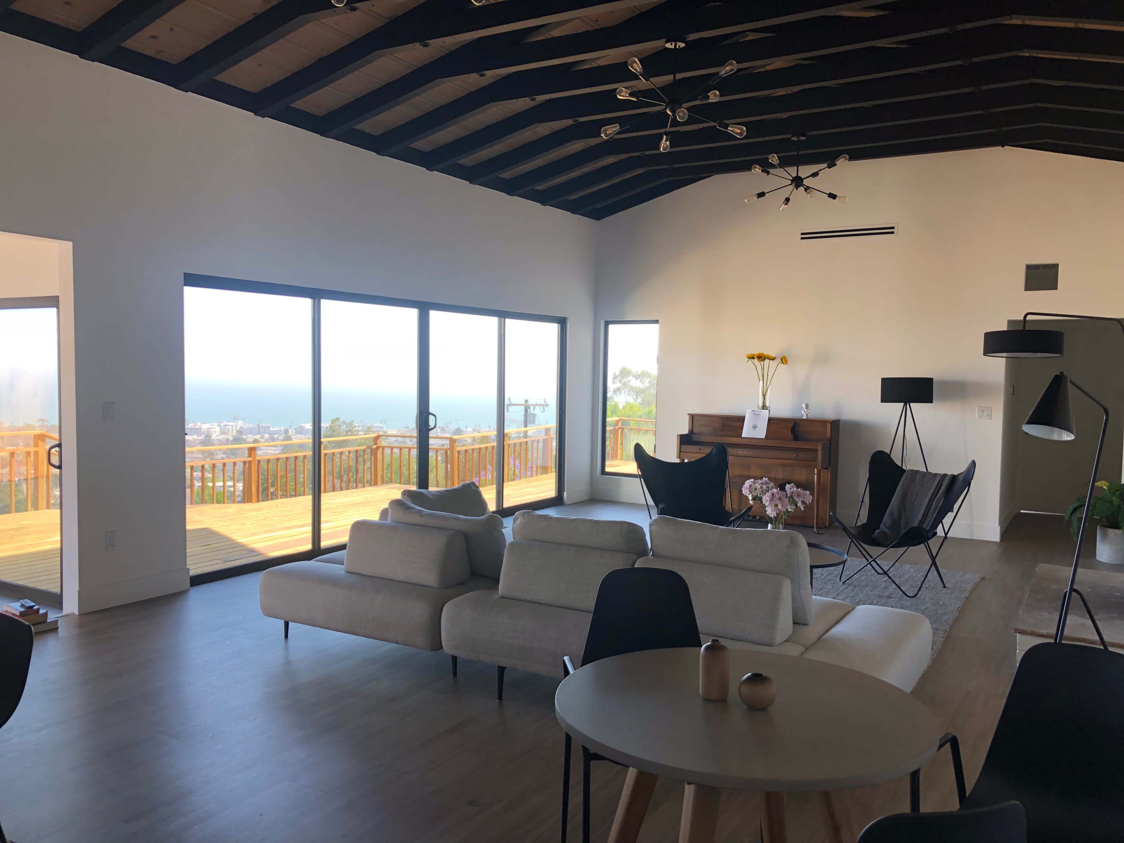 A modern, open-concept living room features large windows overlooking a scenic view, with a minimalist sofa, a round table, and stylish black chairs.
