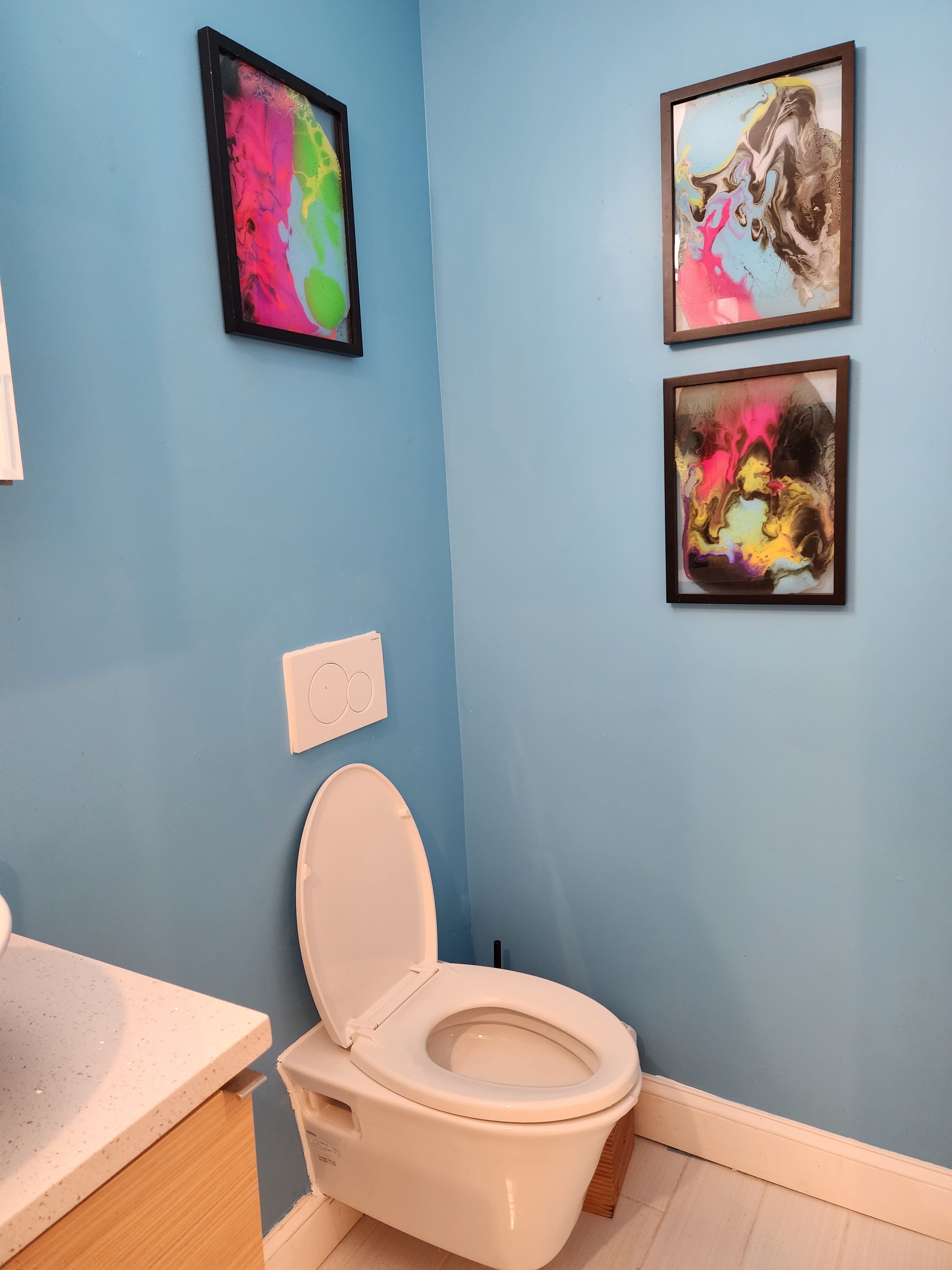 A toilet is positioned against a blue wall featuring two framed abstract art pieces.