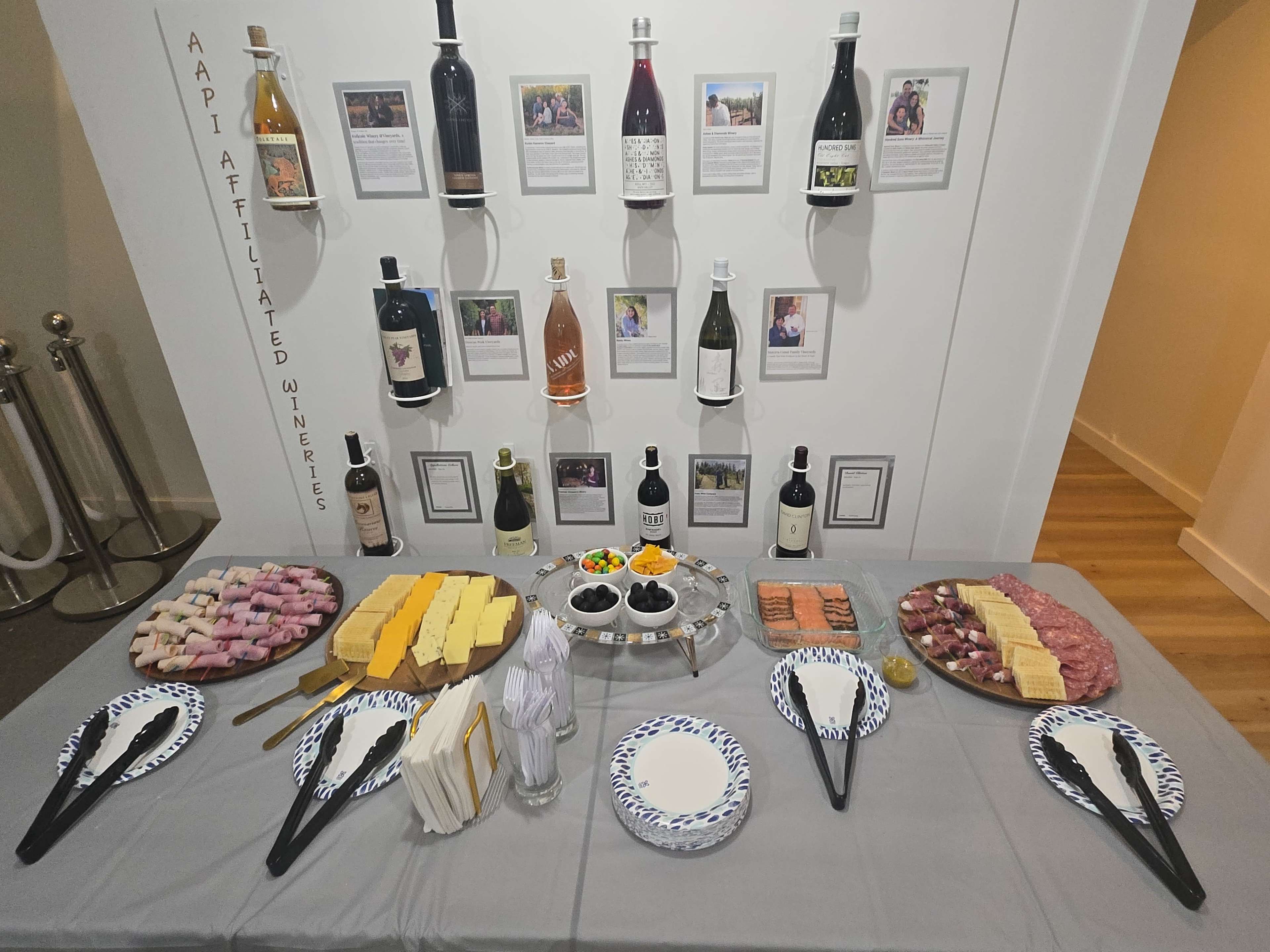 The scene shows a table laden with various food items, including platters of sushi, cheese, and cured meats, in front of a wall displaying bottles of wine and informational cards about AAPI-affiliated wineries.