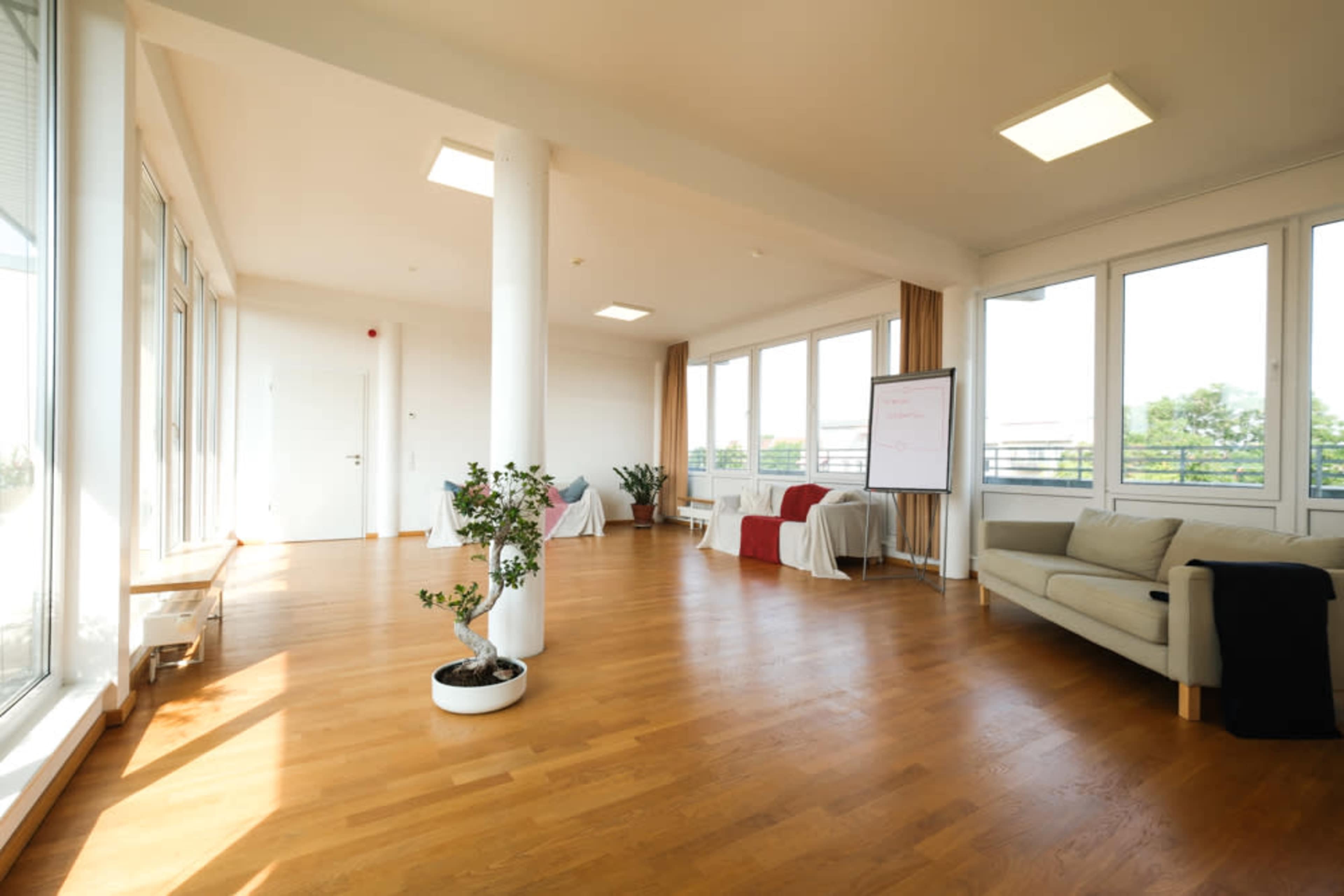 The image shows a brightly lit, spacious room with large windows, hardwood flooring, and a seating area featuring a sofa and a whiteboard.