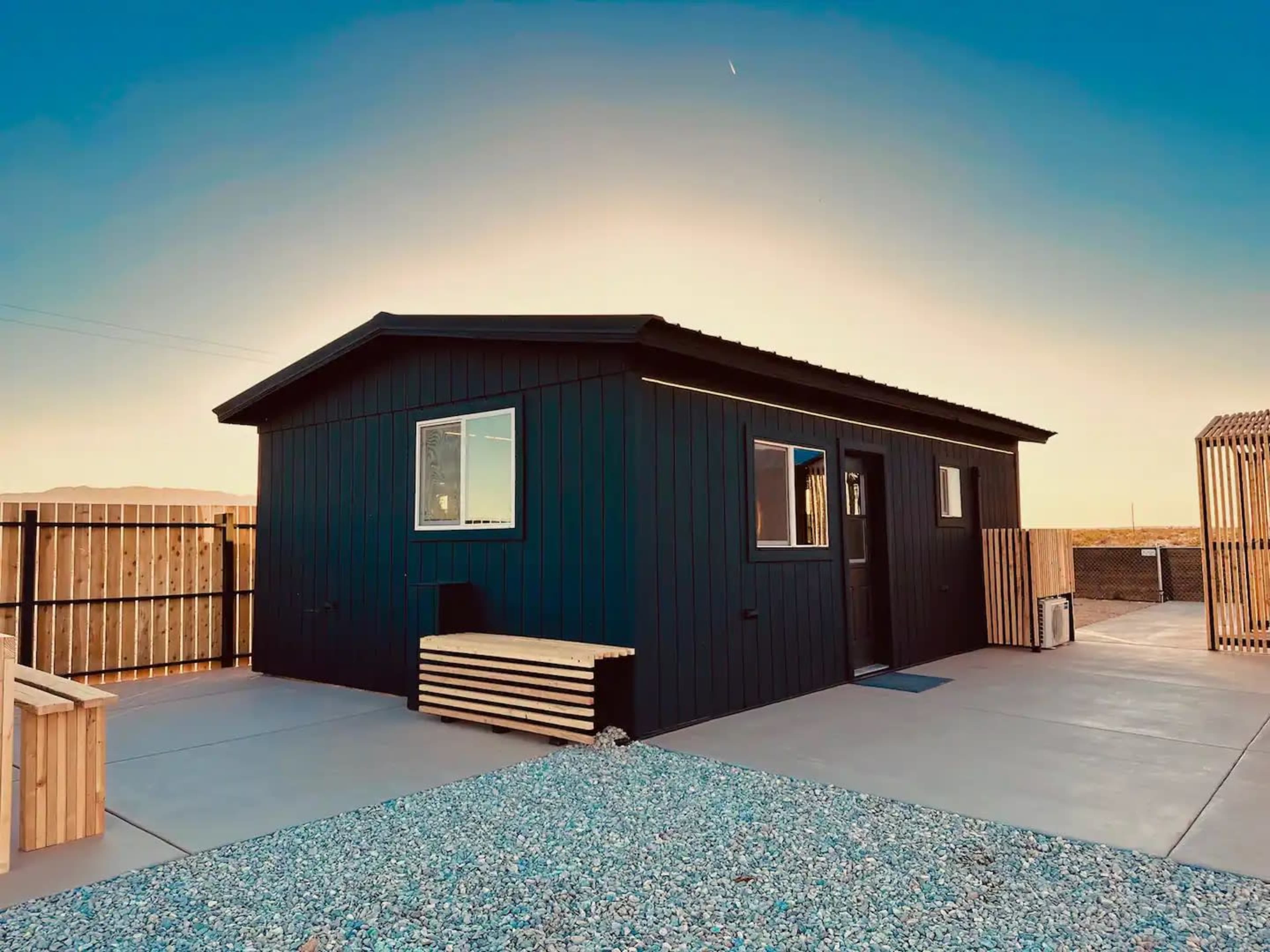 A small black modular house with multiple windows is situated on a gravel lot surrounded by a wooden fence.