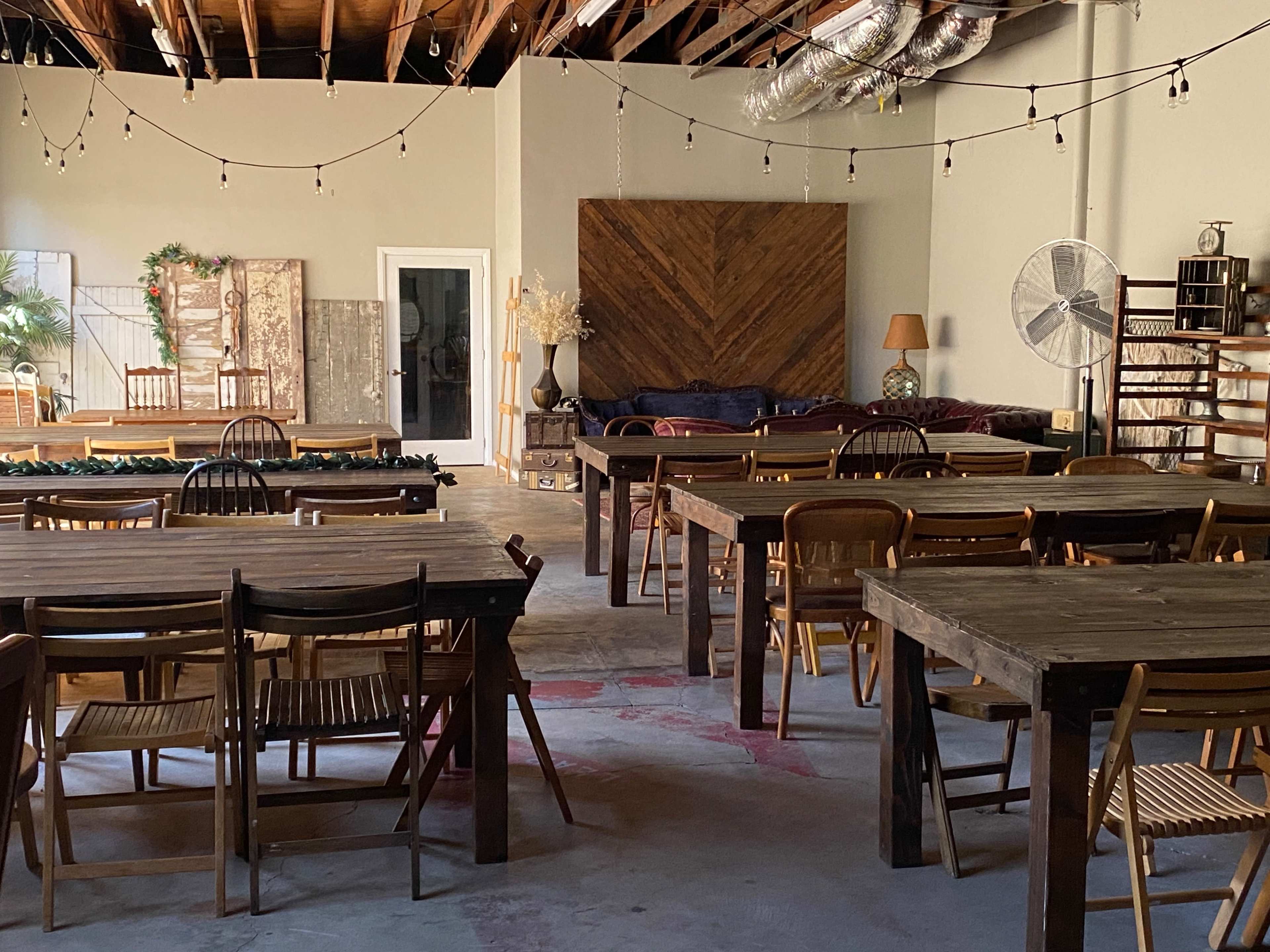 The image shows a spacious, well-lit room with wooden tables and chairs arranged for an event, featuring a cozy seating area and decorative elements on the walls.