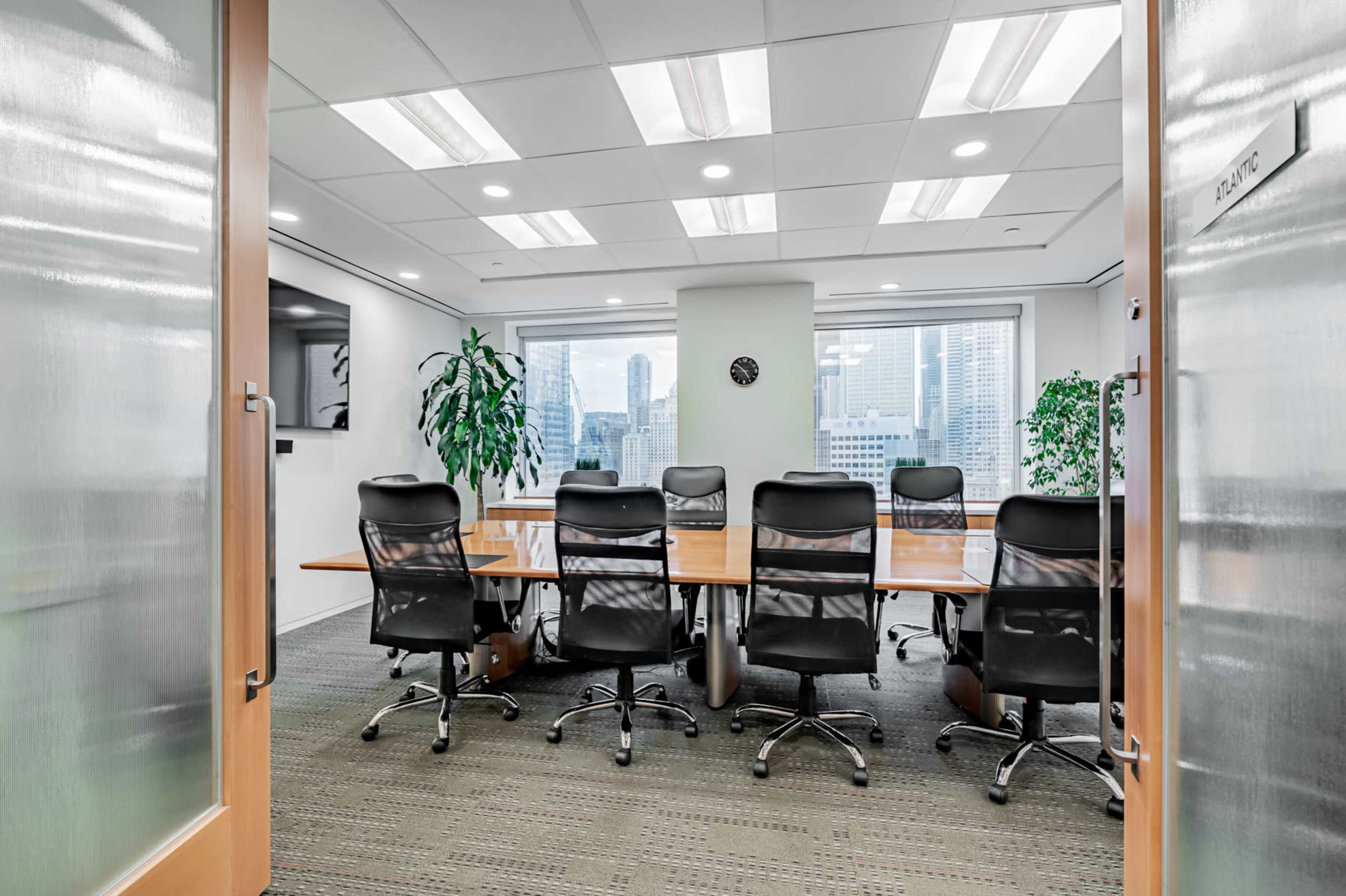 A conference room with a long wooden table and eight black chairs is visible through glass doors, featuring large windows that overlook a city skyline.