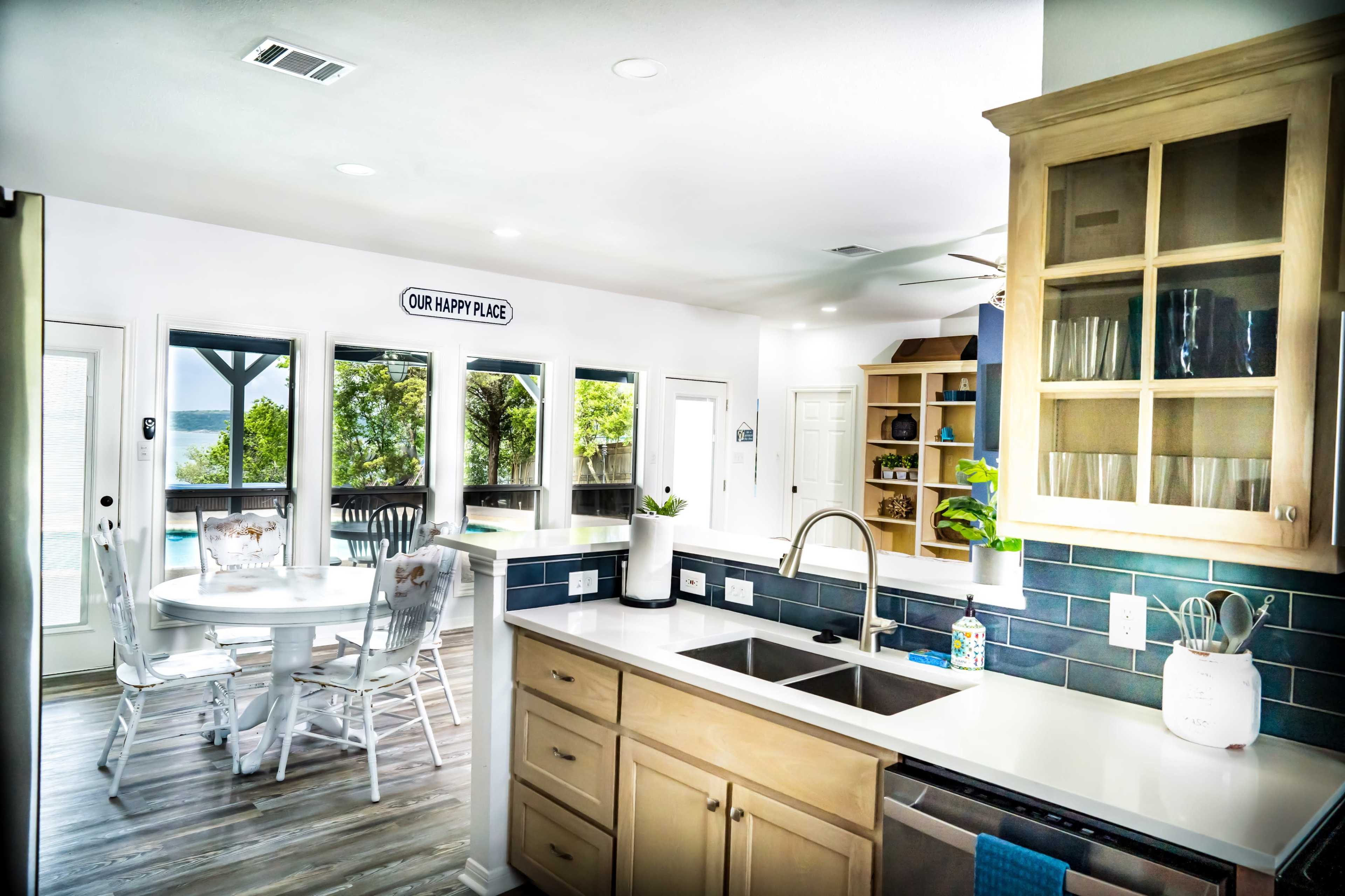 A modern kitchen with light wood cabinets, a white countertop, and a dining area featuring a circular table and chairs, alongside large windows that let in natural light.