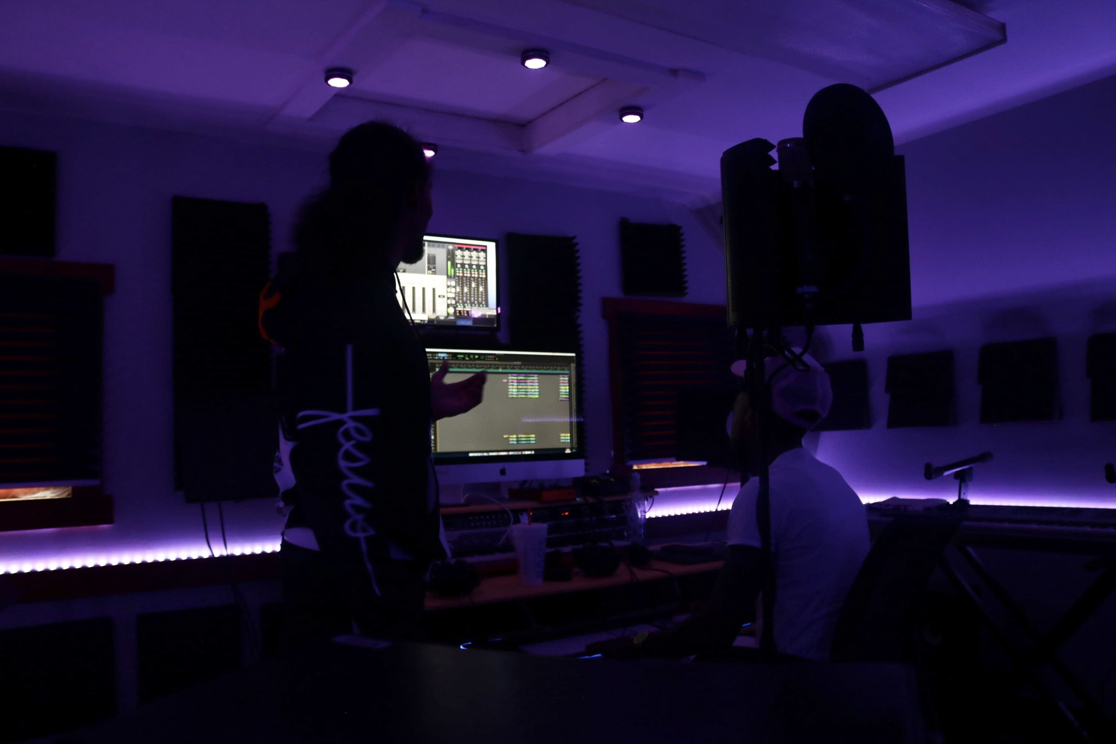 Two individuals are engaged in a conversation in a dimly lit music studio with computers and sound equipment.