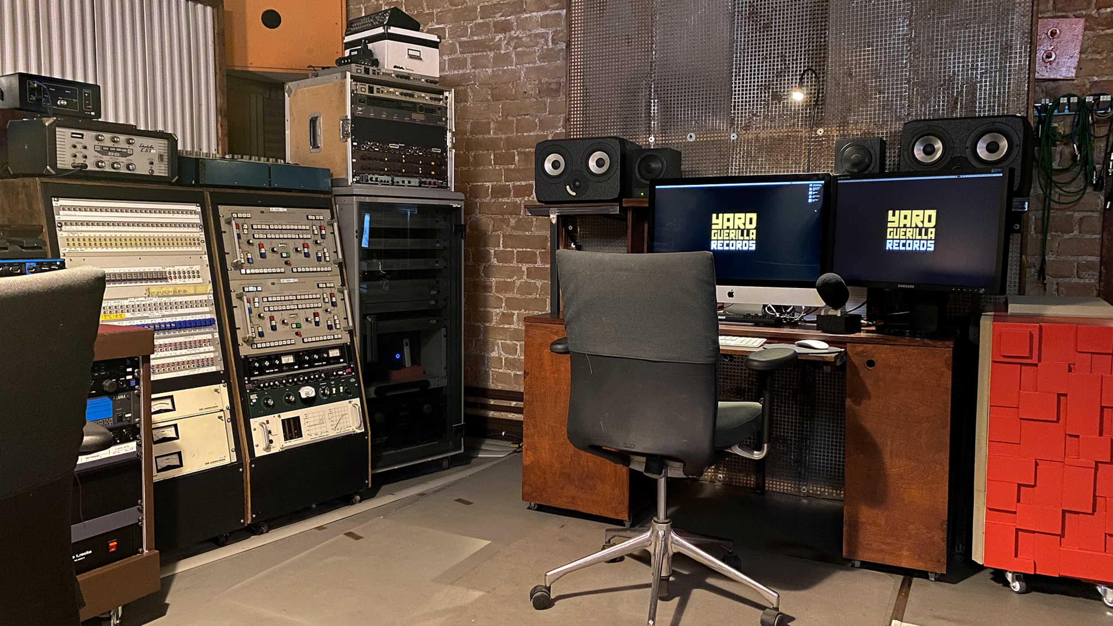The image shows a sound studio featuring a recording console, audio equipment racks, and dual computer monitors set up at a desk with a swivel chair.