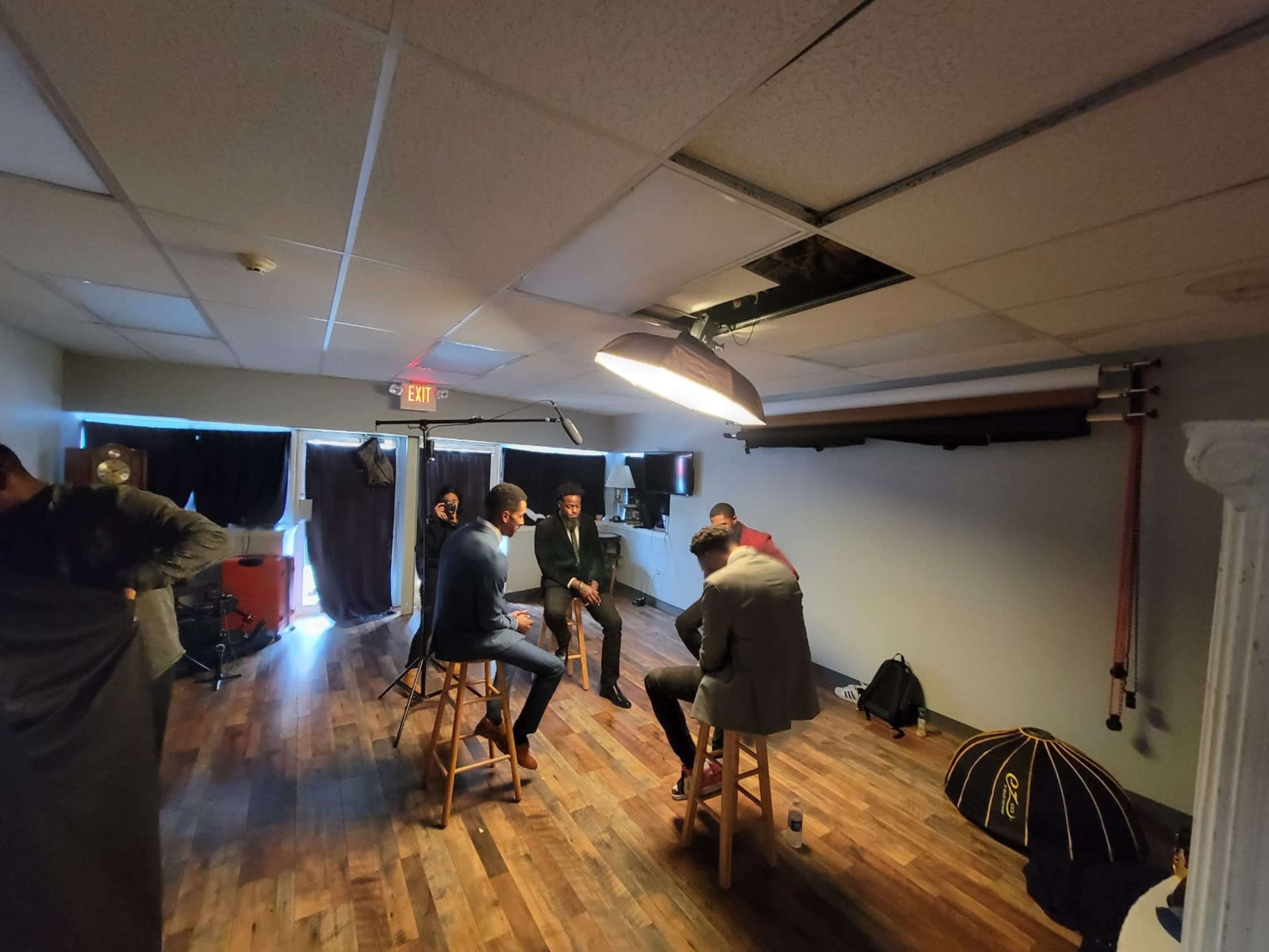 A group of five individuals sits in a studio space with wooden flooring, engaged in a discussion while surrounded by photography equipment.