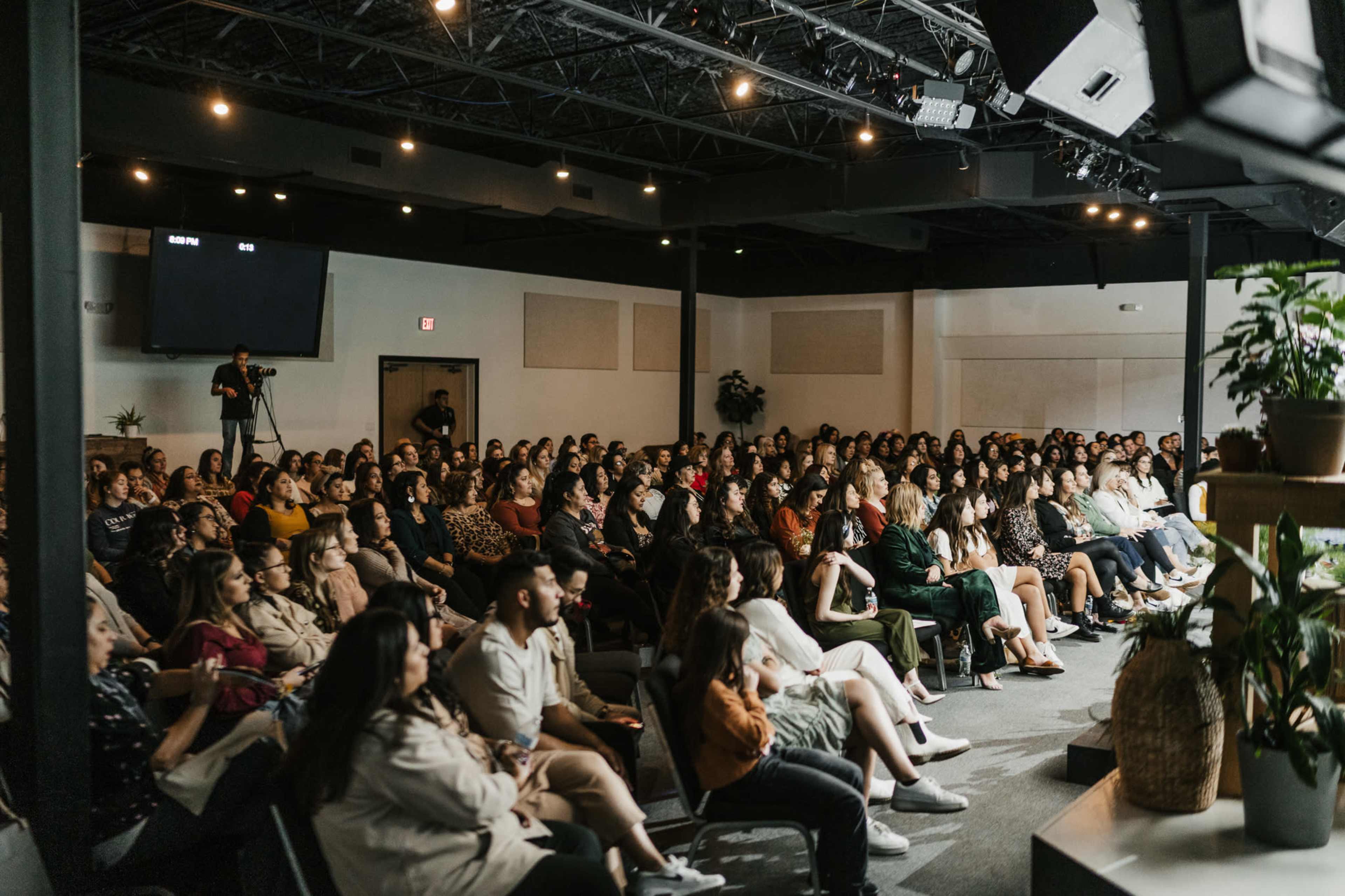 A large audience is seated in a modern lecture space, attentively facing a stage with a speaker and a screen.