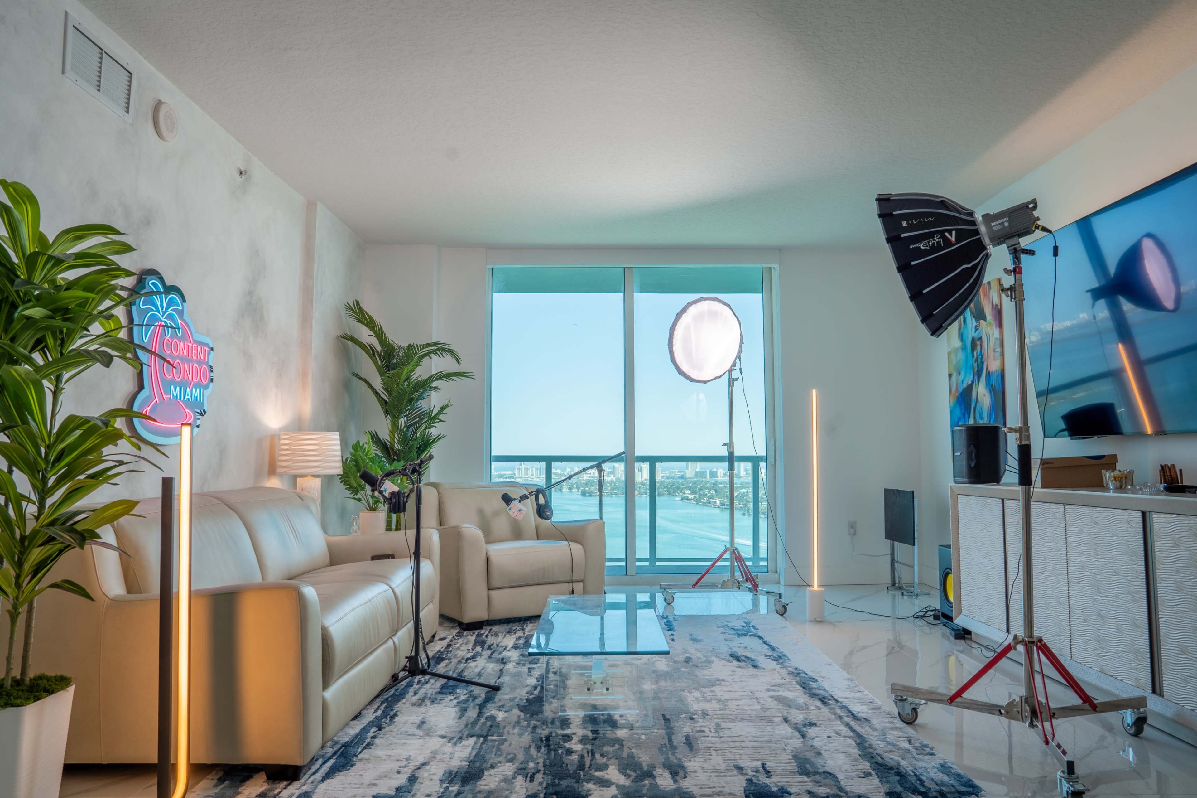The image shows a modern living room with two beige sofas, a glass coffee table, and large windows overlooking a waterfront view, accompanied by studio lighting equipment.