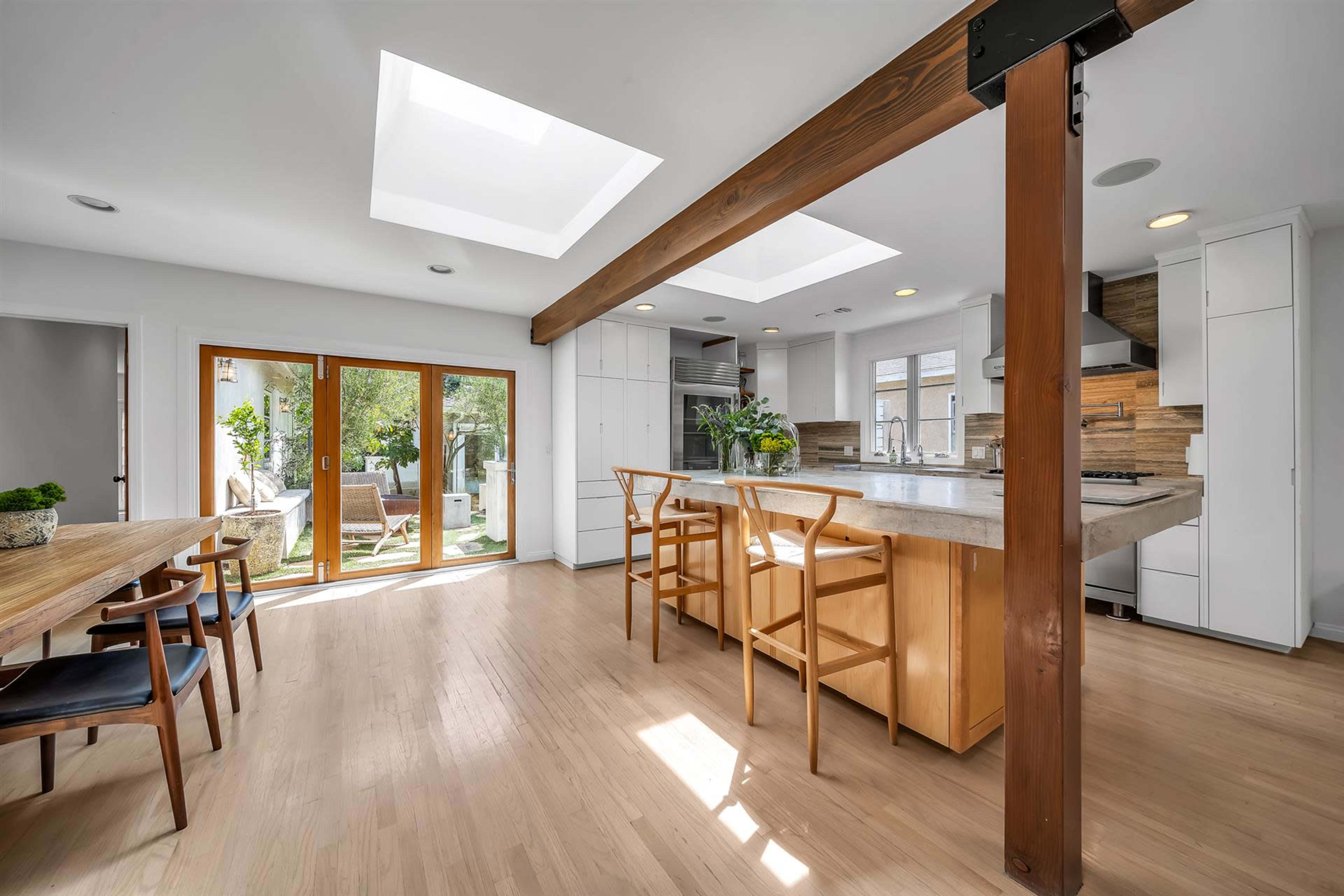 The image shows a modern kitchen and dining area featuring a central island, skylights, and large windows opening to a patio.