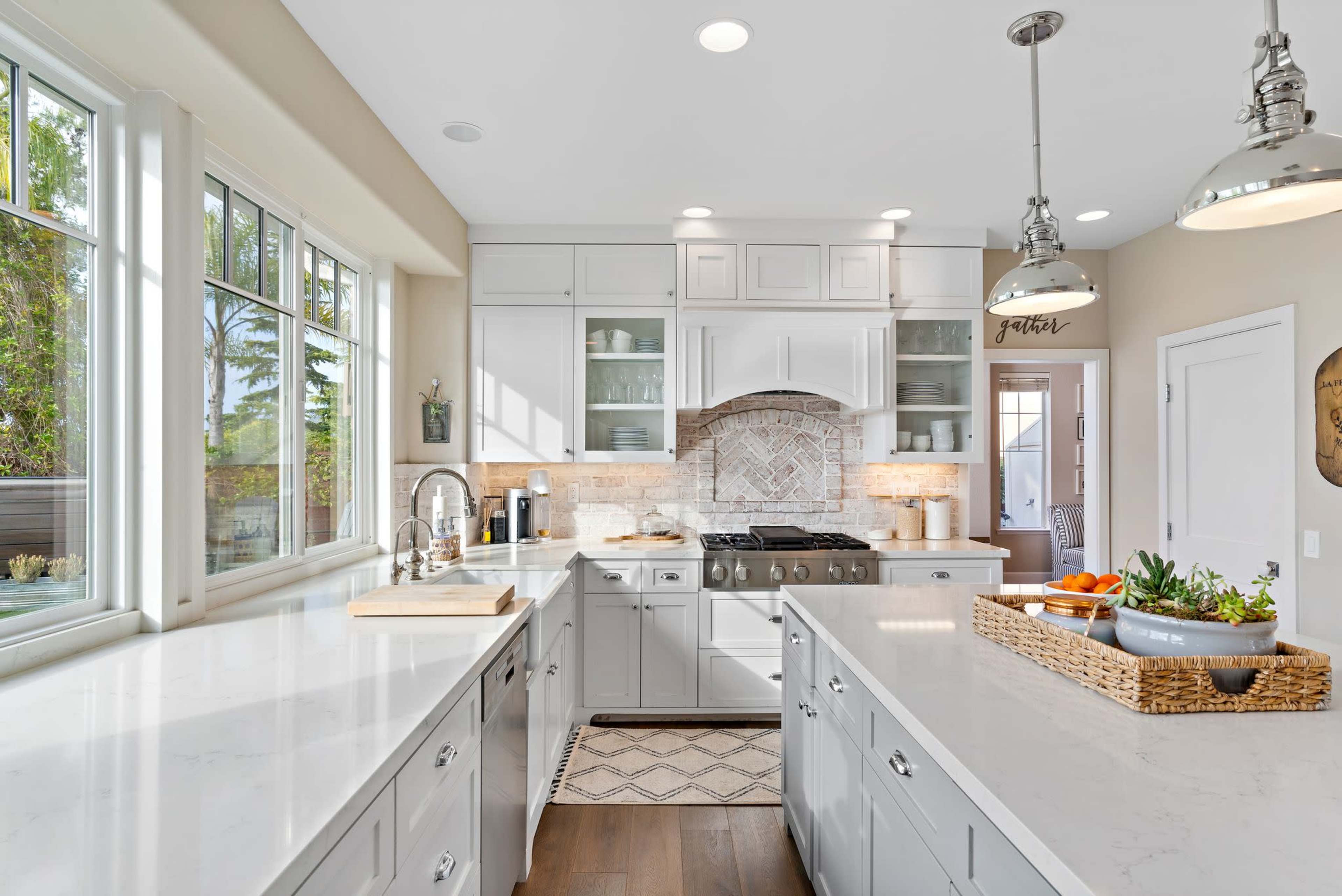 A modern kitchen features white cabinetry, a marble countertop, and stainless steel appliances, with natural light streaming in through large windows.