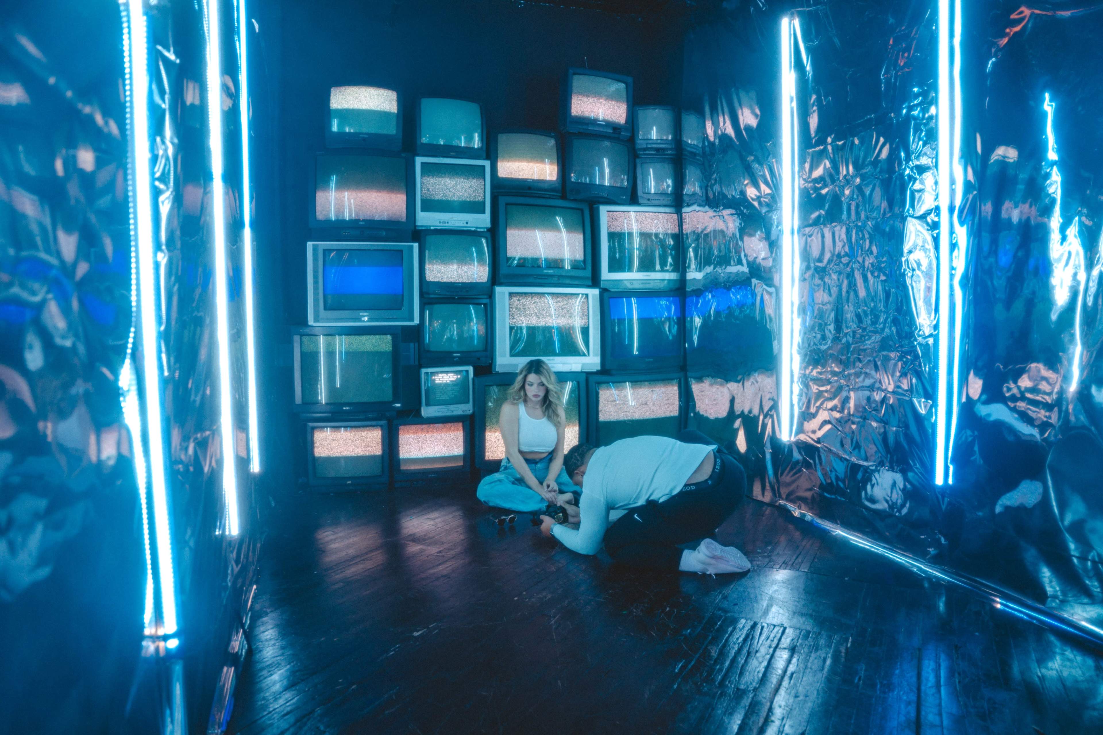 A woman sits on the floor in a room illuminated by blue neon lights, surrounded by vintage televisions displaying static.