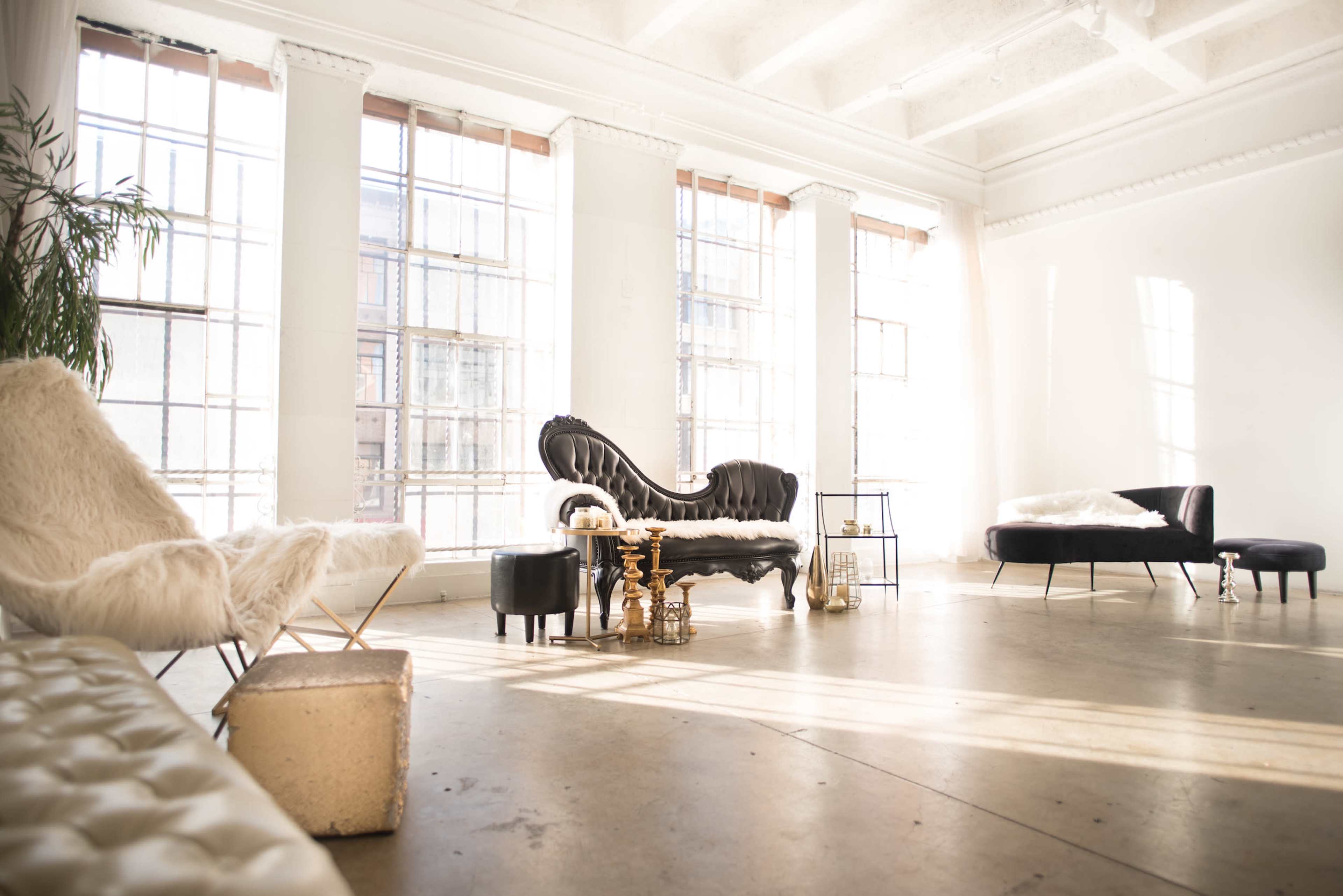 A bright, spacious room with large windows, featuring a mix of modern and vintage furniture, including a black sofa and a white armchair.