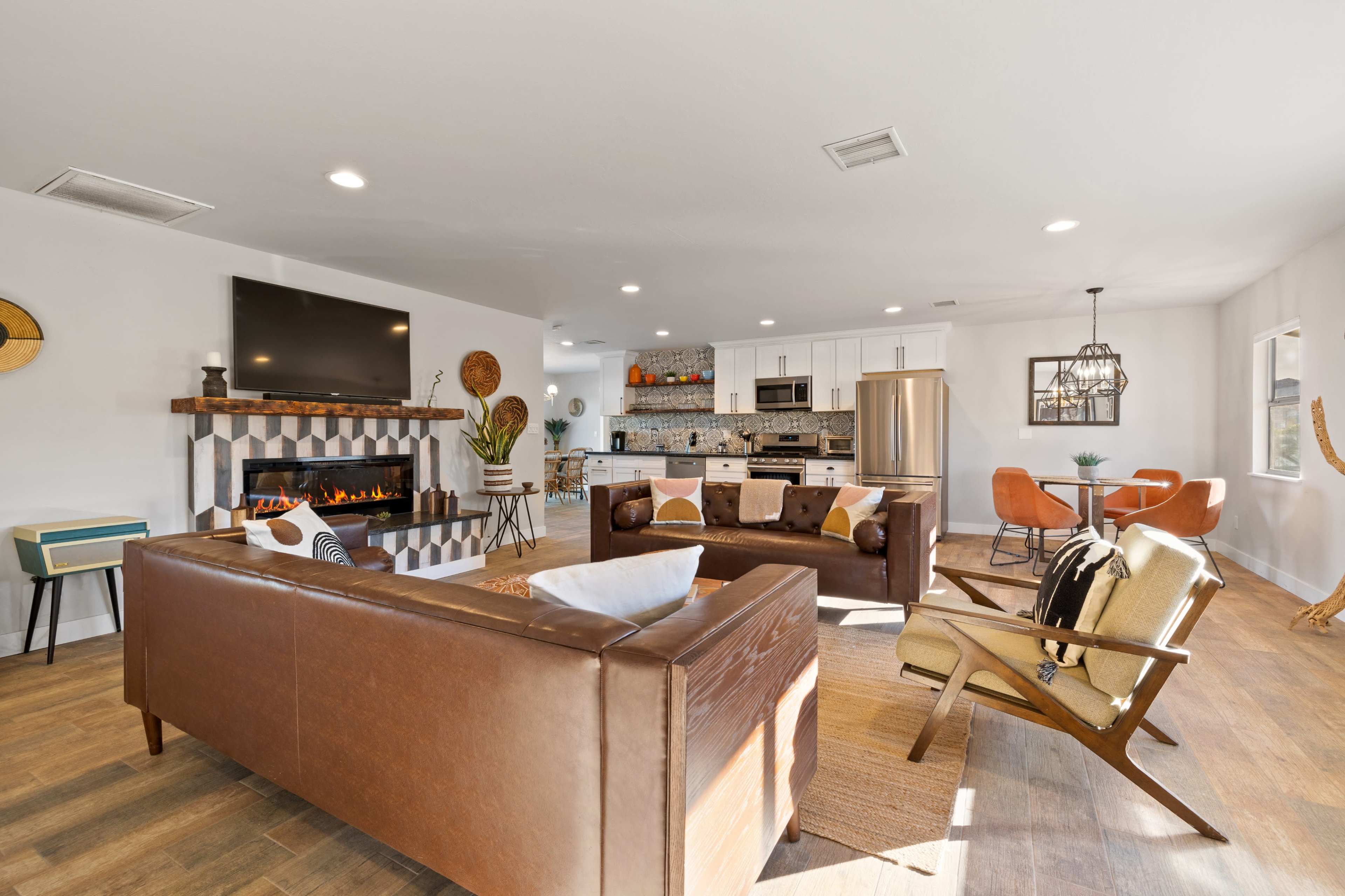 The image shows a modern living room with a leather sofa, a fireplace, and an open layout connecting to a dining area with a round table and orange chairs.