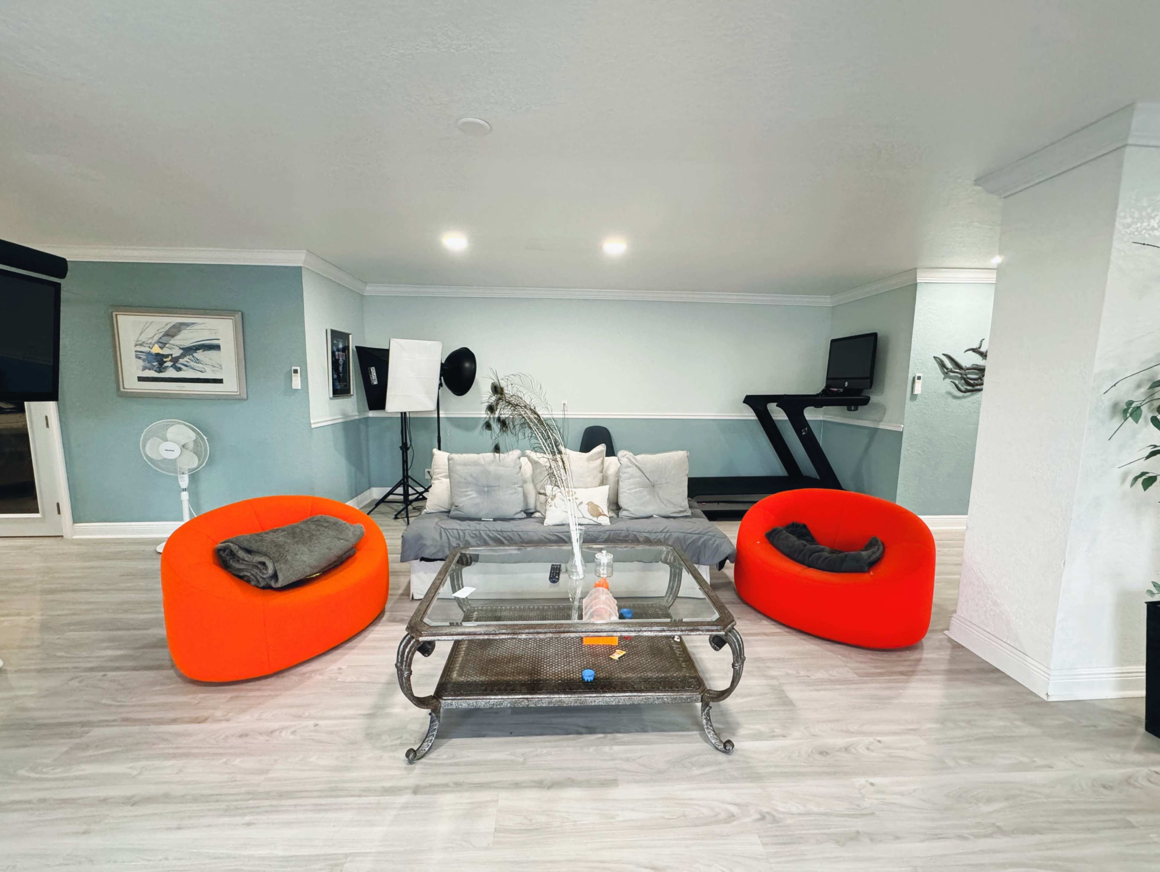 A modern living area features two bright red chairs, a glass coffee table, and a treadmill, with light-colored walls and a fan in the background.