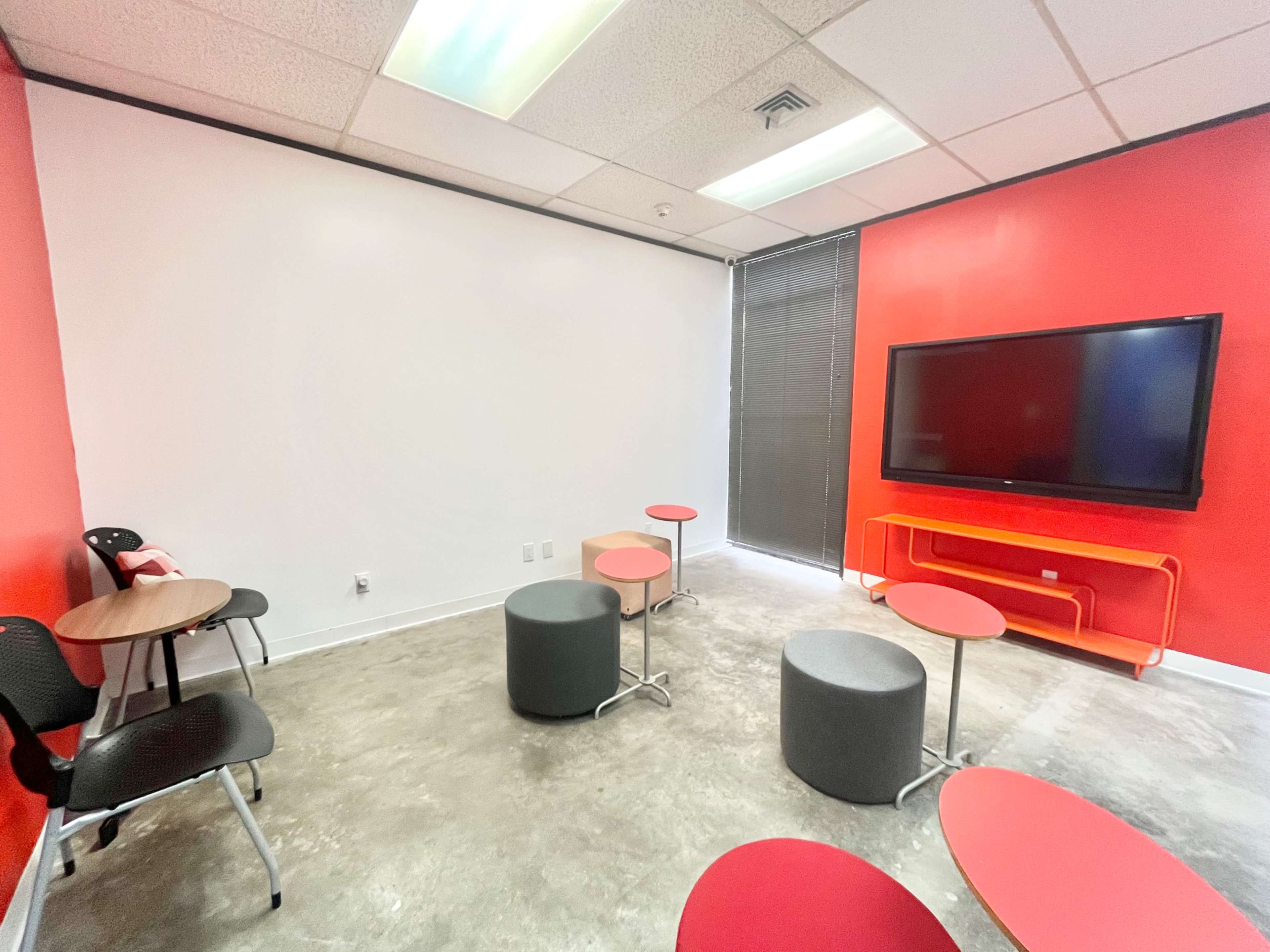 The image shows a small meeting room with red walls, a large TV mounted on one wall, and several round and oval tables with chairs.