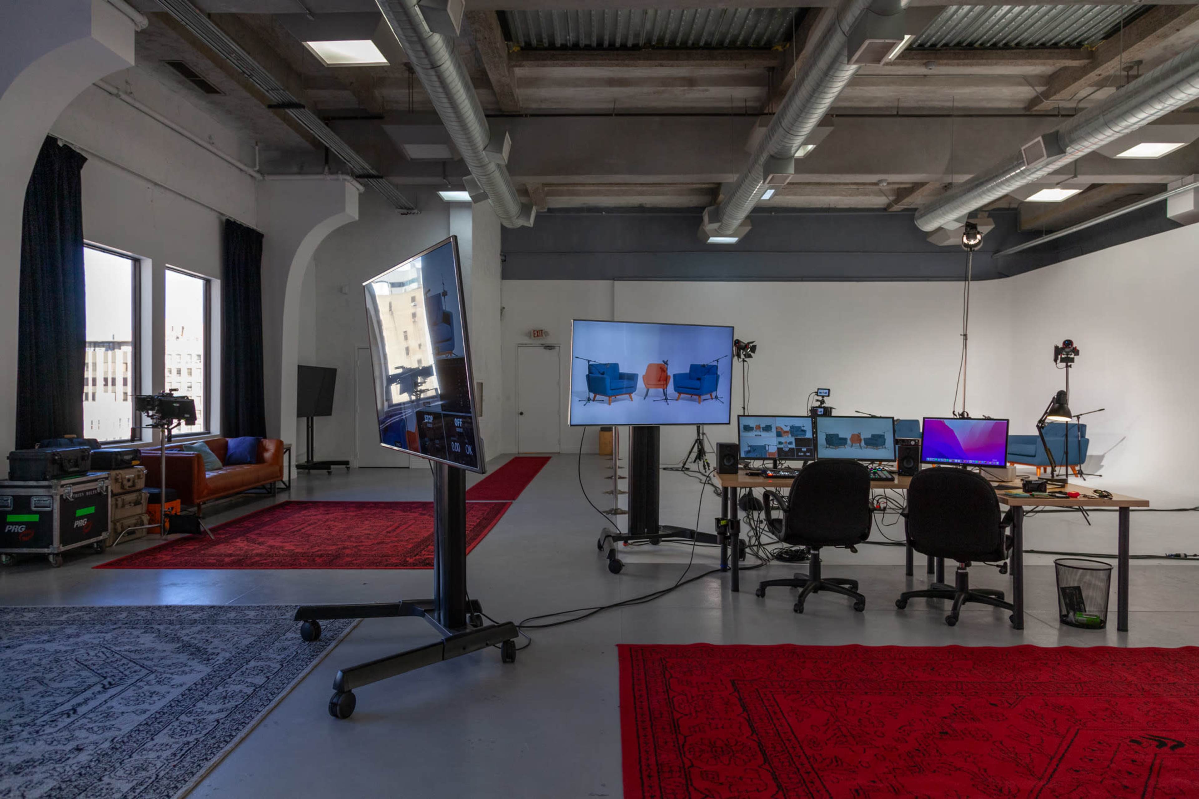 An empty studio features a blue sofa, two large screens displaying images, and a workspace with computers and cameras on red rugs.