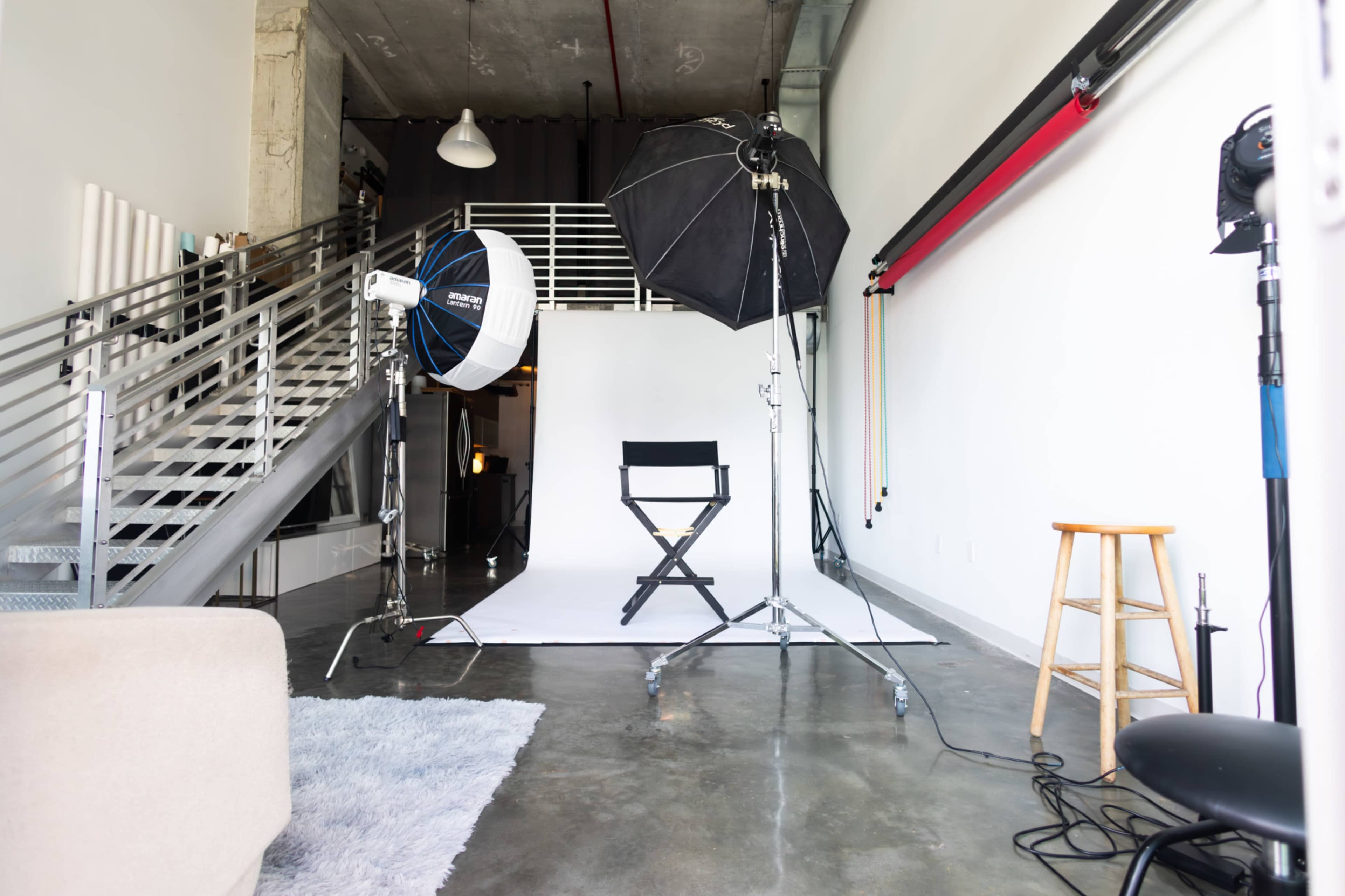 The image shows a photography studio featuring a white backdrop, a director's chair, and lighting equipment arranged on a polished concrete floor.