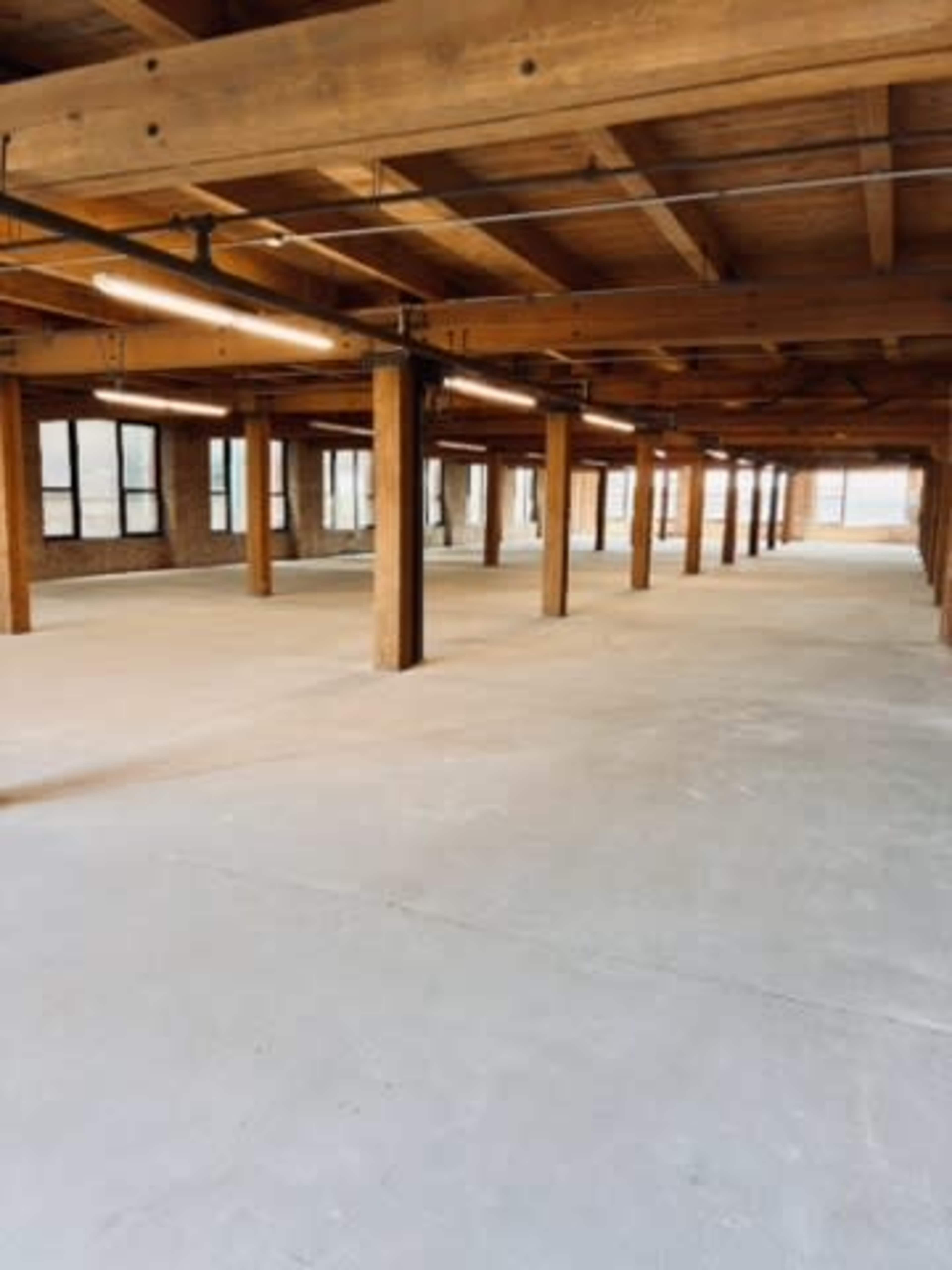 The image shows an empty interior space with wooden beams and columns, large windows, and a concrete floor.