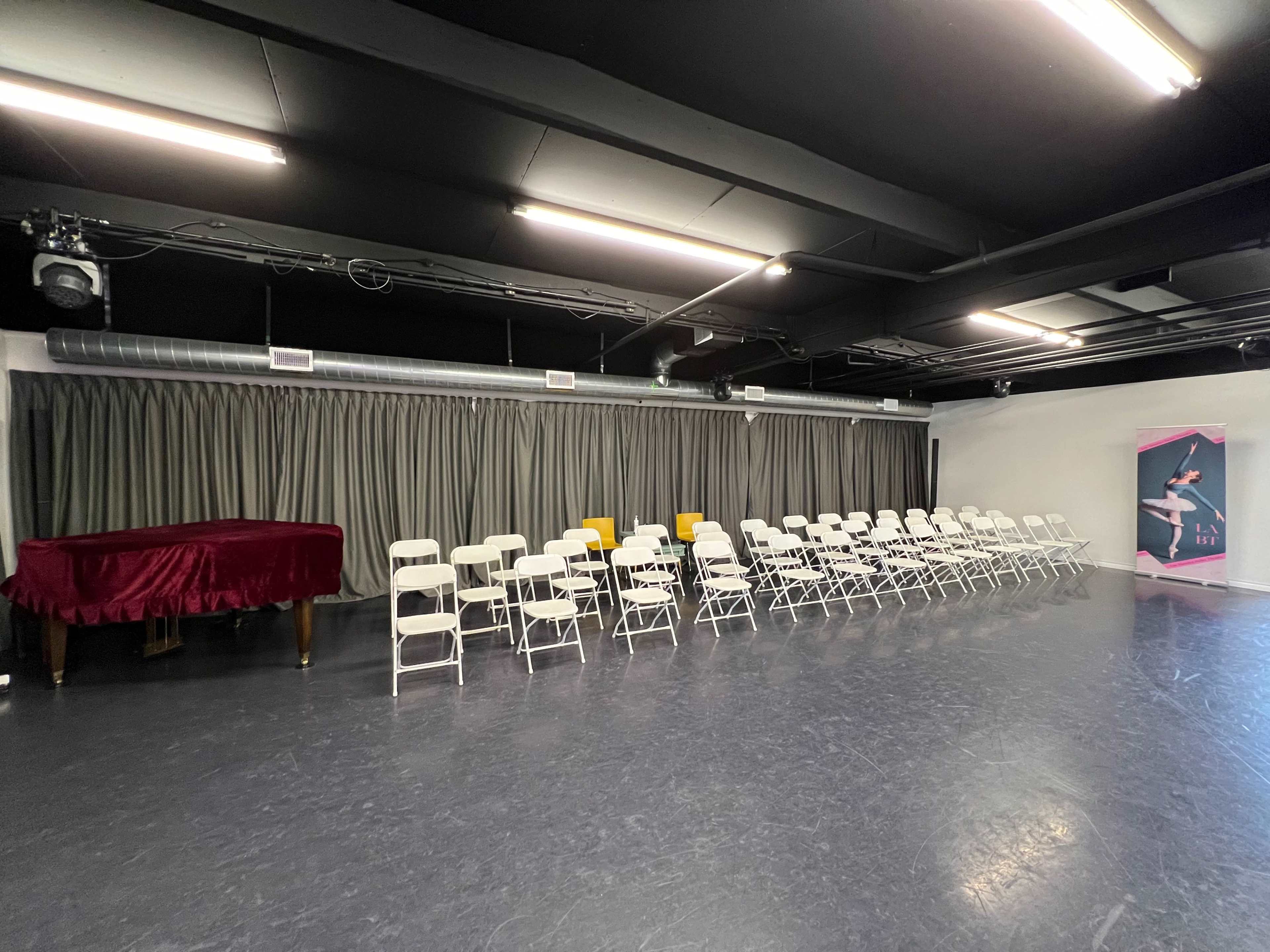 The image shows a dance studio with rows of white chairs, a red piano, and a curtain along one wall.