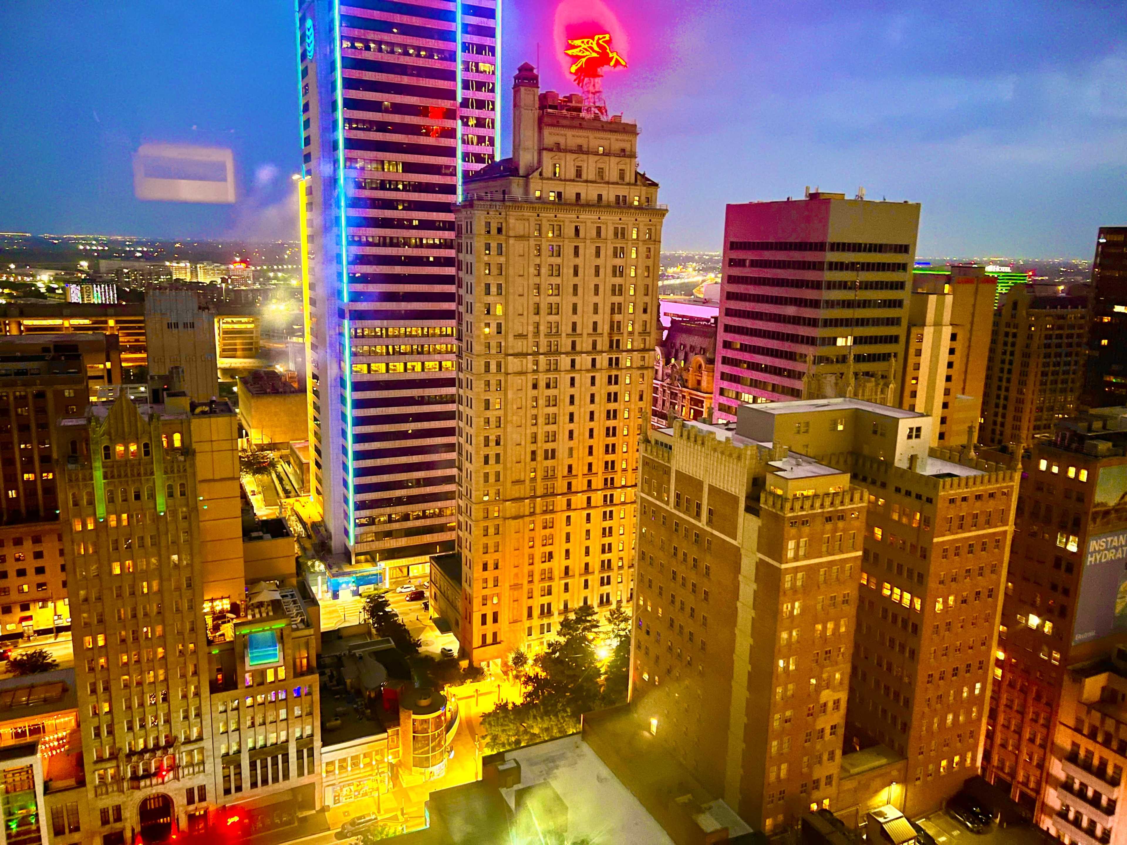 A city skyline at dusk showcases brightly lit buildings, with a prominent structure topped by a neon sign of a flying red bird.