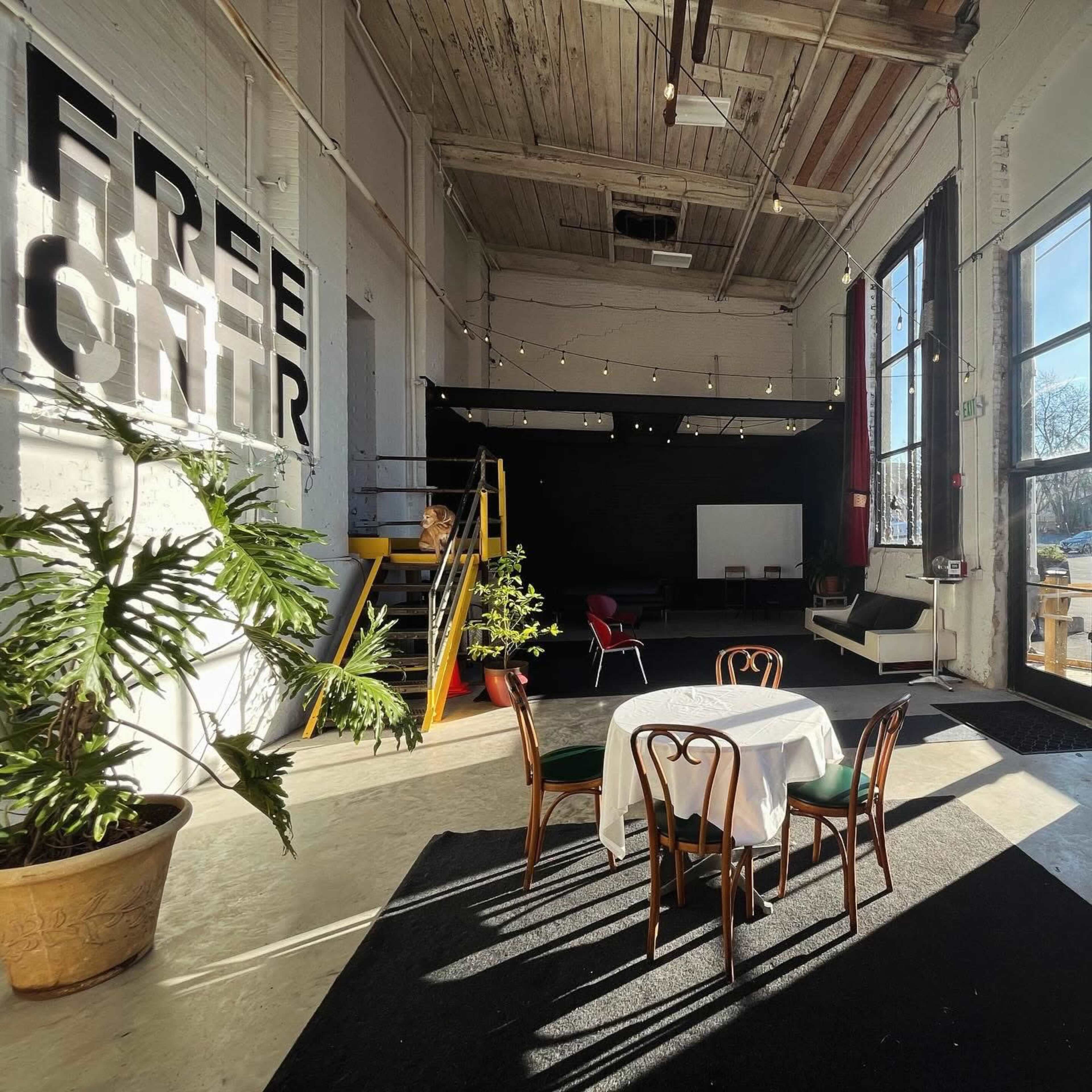 The image shows a spacious interior with high ceilings, featuring a small round table with four chairs, a potted plant, and a stage area in the background.