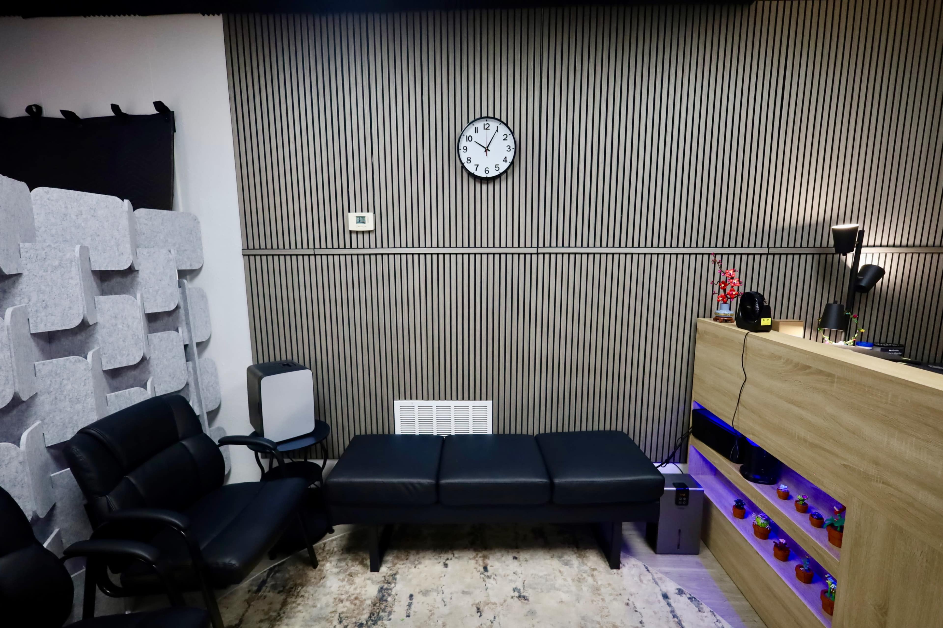The image shows a modern, minimalistic room featuring a black sofa, a wooden shelving unit with colored lights, a wall clock, and sound-absorbing panels.