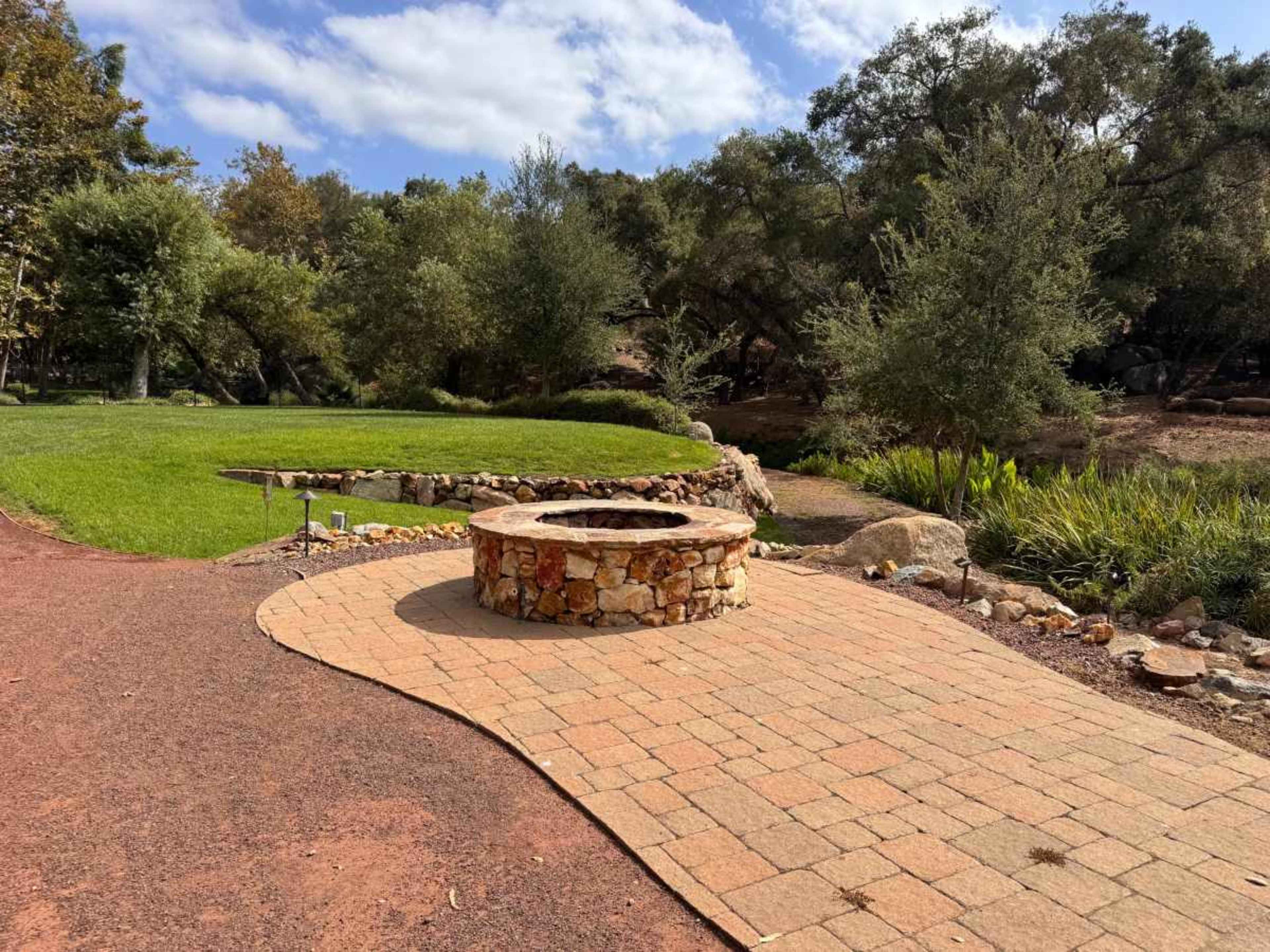 The image shows a circular stone fire pit surrounded by a paved pathway, with grass and trees in the background.