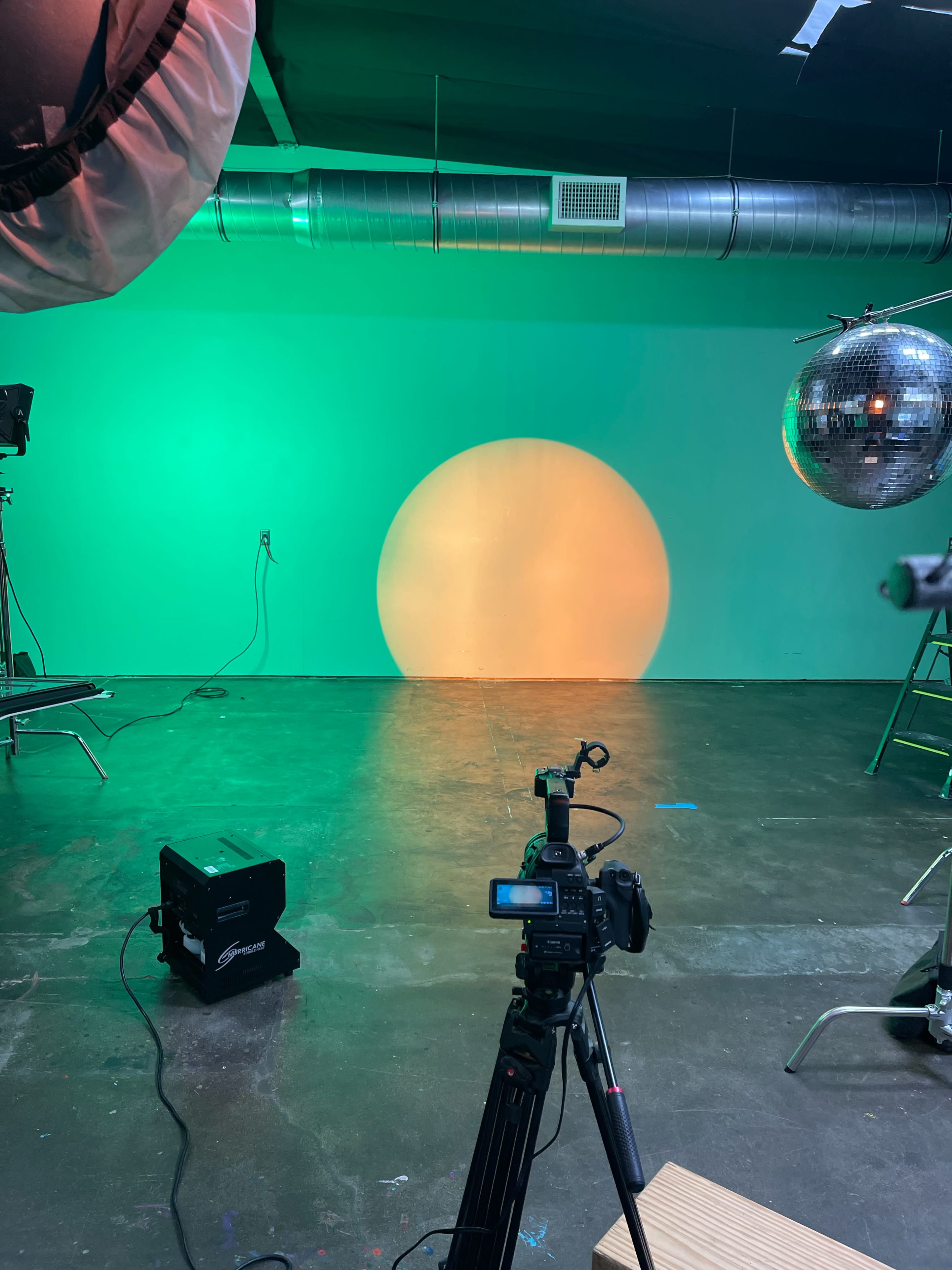 A camera is set up in a studio with colored lights casting a circular shadow on a white wall, alongside a disco ball and studio equipment.