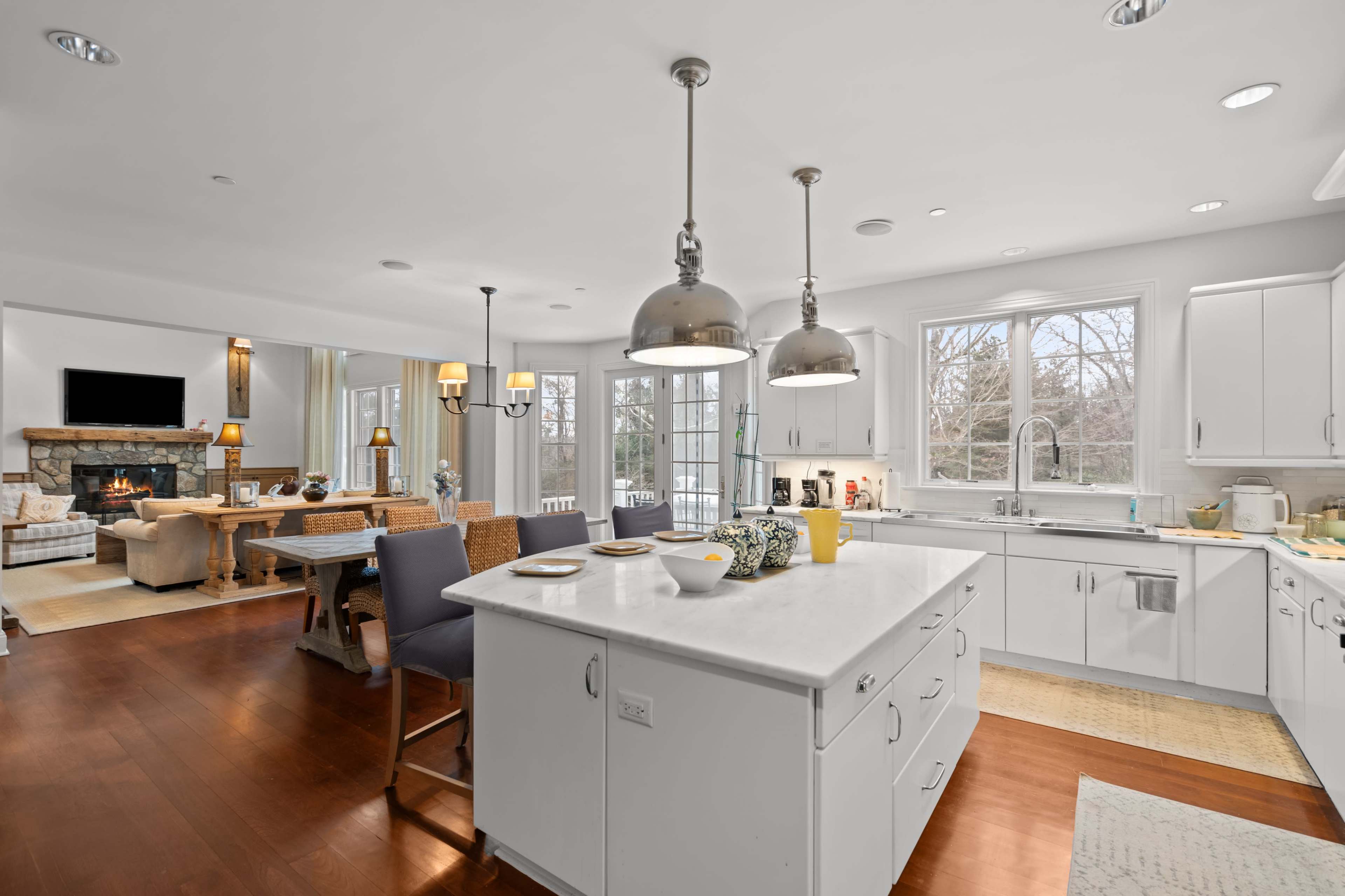 The image shows a modern kitchen with an island, pendant lighting, and an open layout connecting to a living area featuring a stone fireplace.
