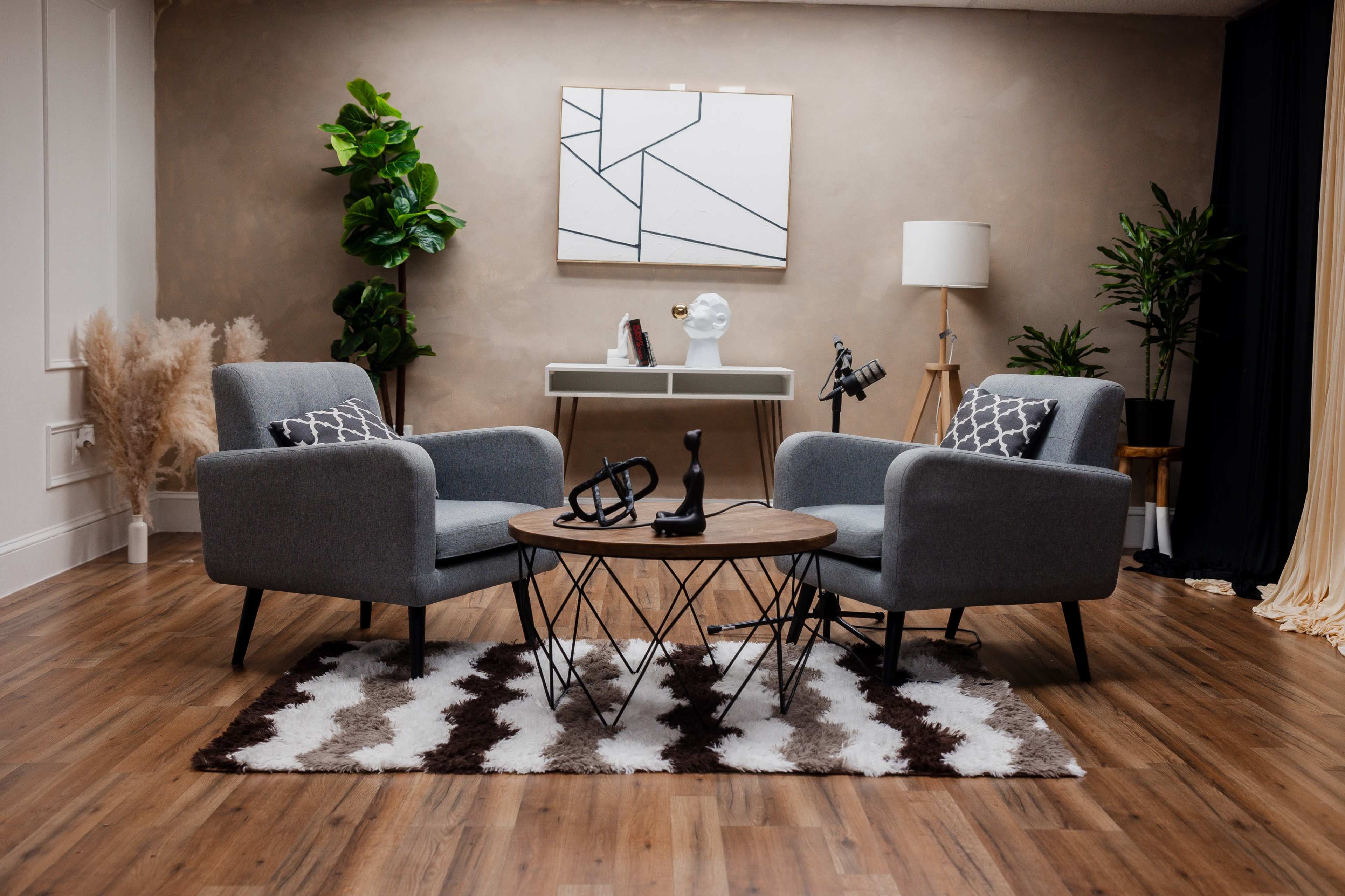 A modern living room features two gray armchairs facing a circular coffee table on a patterned rug, with plants and decorative objects arranged throughout the space.