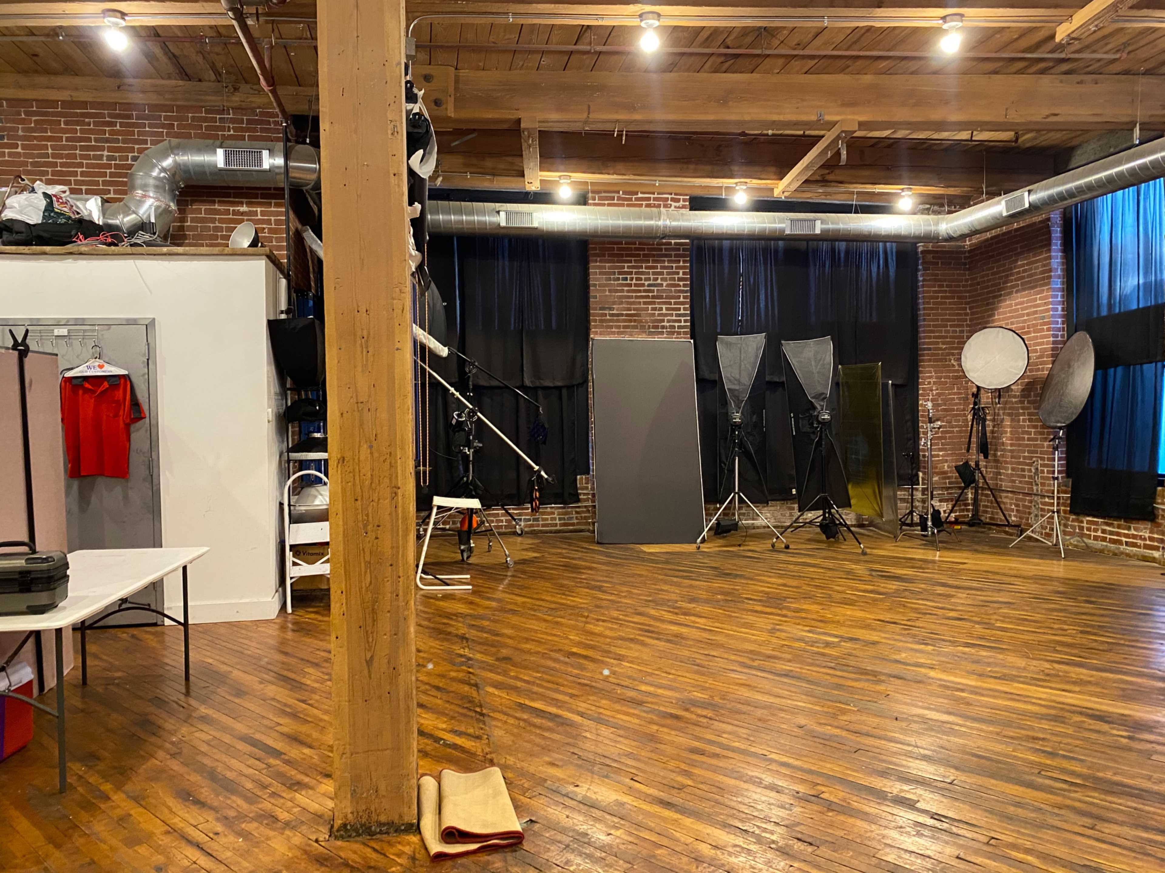 A spacious photo studio with wooden floors, exposed brick walls, and various lighting equipment arranged throughout the room.
