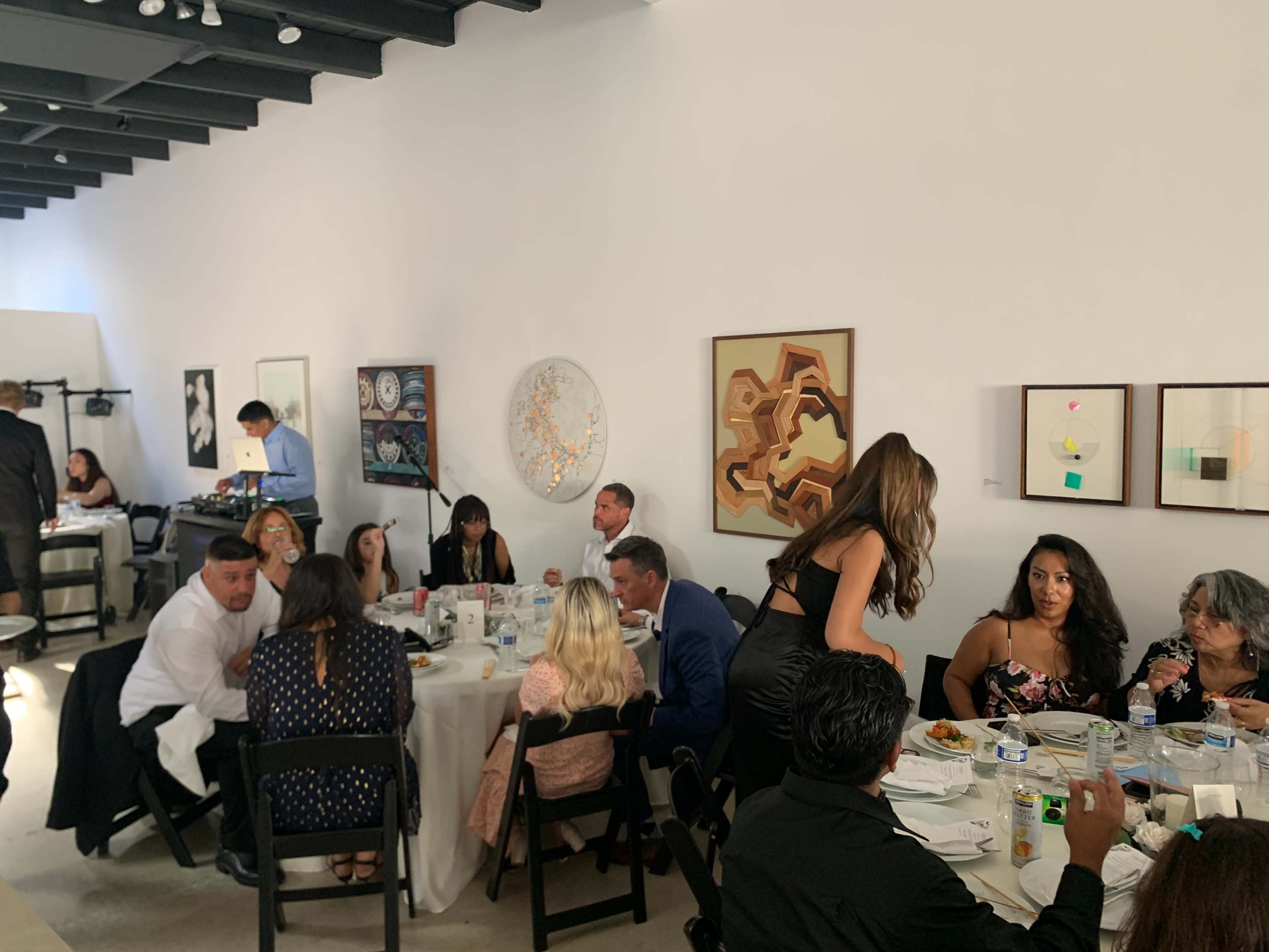 A group of people is seated at elegantly set tables in a well-lit room adorned with various artworks on the walls.