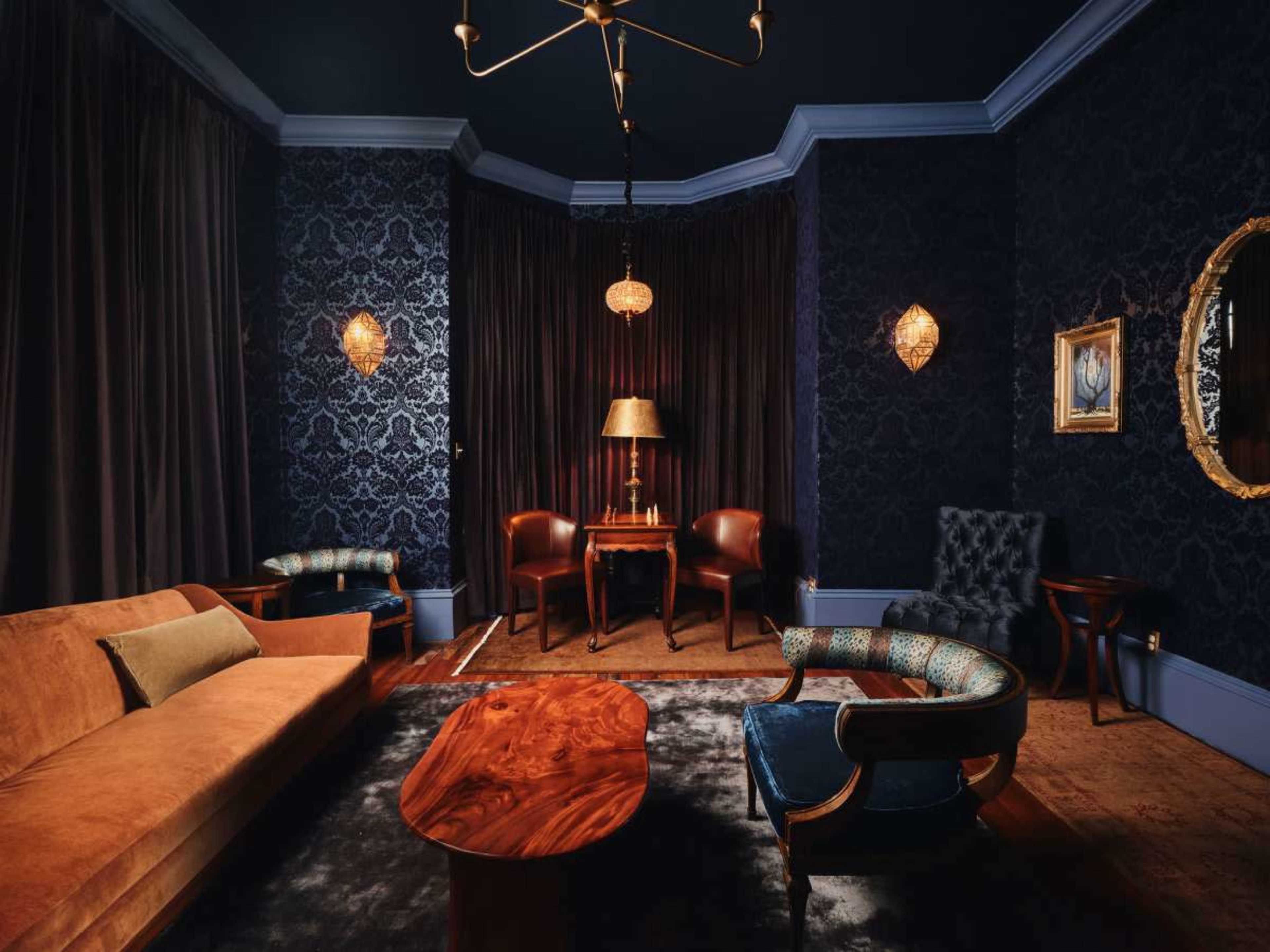 The image shows a dimly lit sitting room featuring dark blue walls, a velvet sofa, two chairs, and a wooden coffee table under a chandelier.